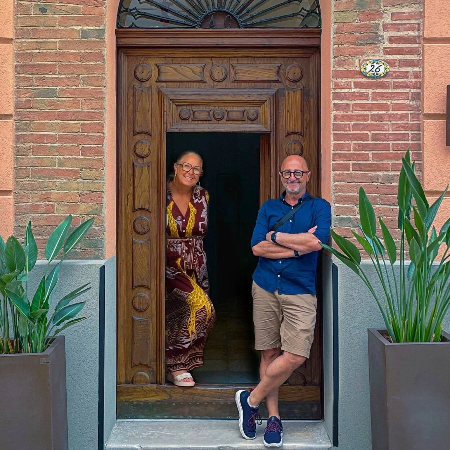 A woman and a man standing at a doorway. The woman is inside the doorway, smiling, wearing glasses and a colorful dress. The man is outside, crossing his arms, wearing glasses, a blue shirt, khaki shorts, and sneakers, leaning casually against the doorway frame. The door is made of wood, and there are large potted plants on either side. The building has a brick exterior with a house number 26 on the wall to the right.