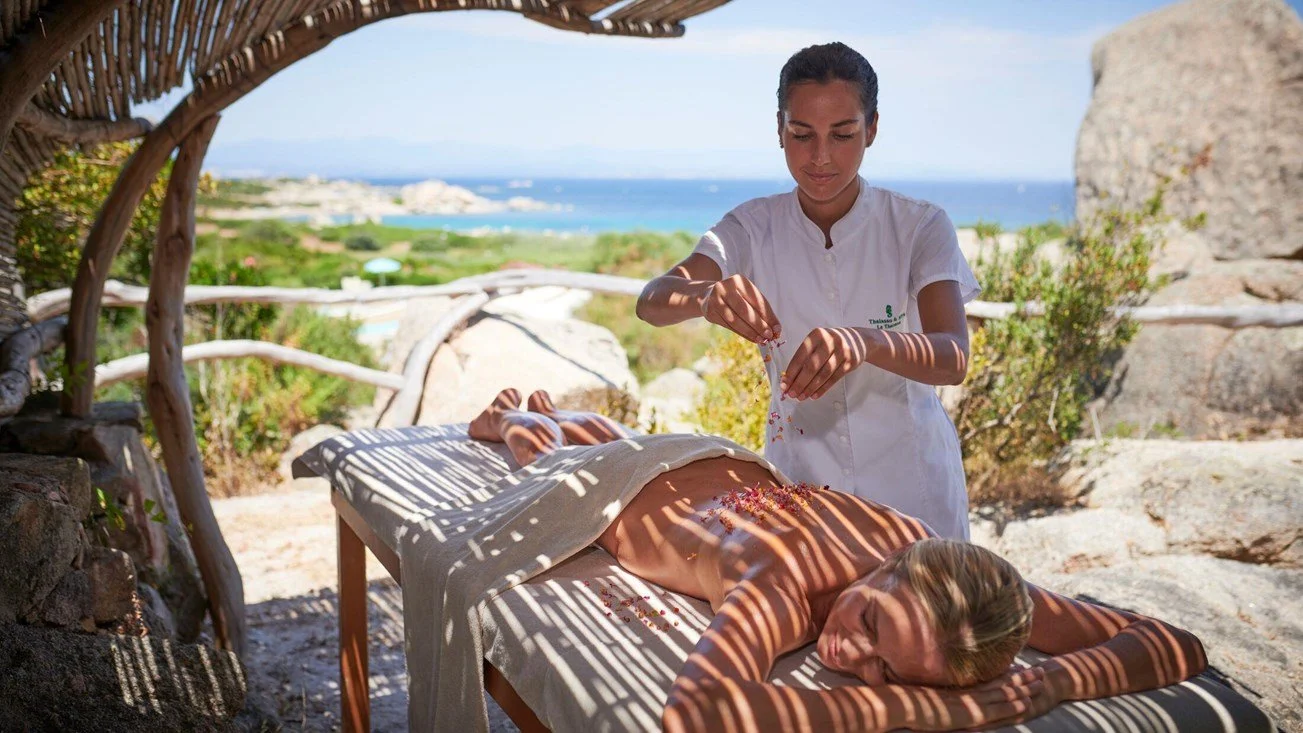 A woman receives an outdoor massage on a lounge bed under a shaded structure overlooking a scenic beach and ocean. A massage therapist applies treatment, and the woman is lying face down with a blanket covering her lower body.