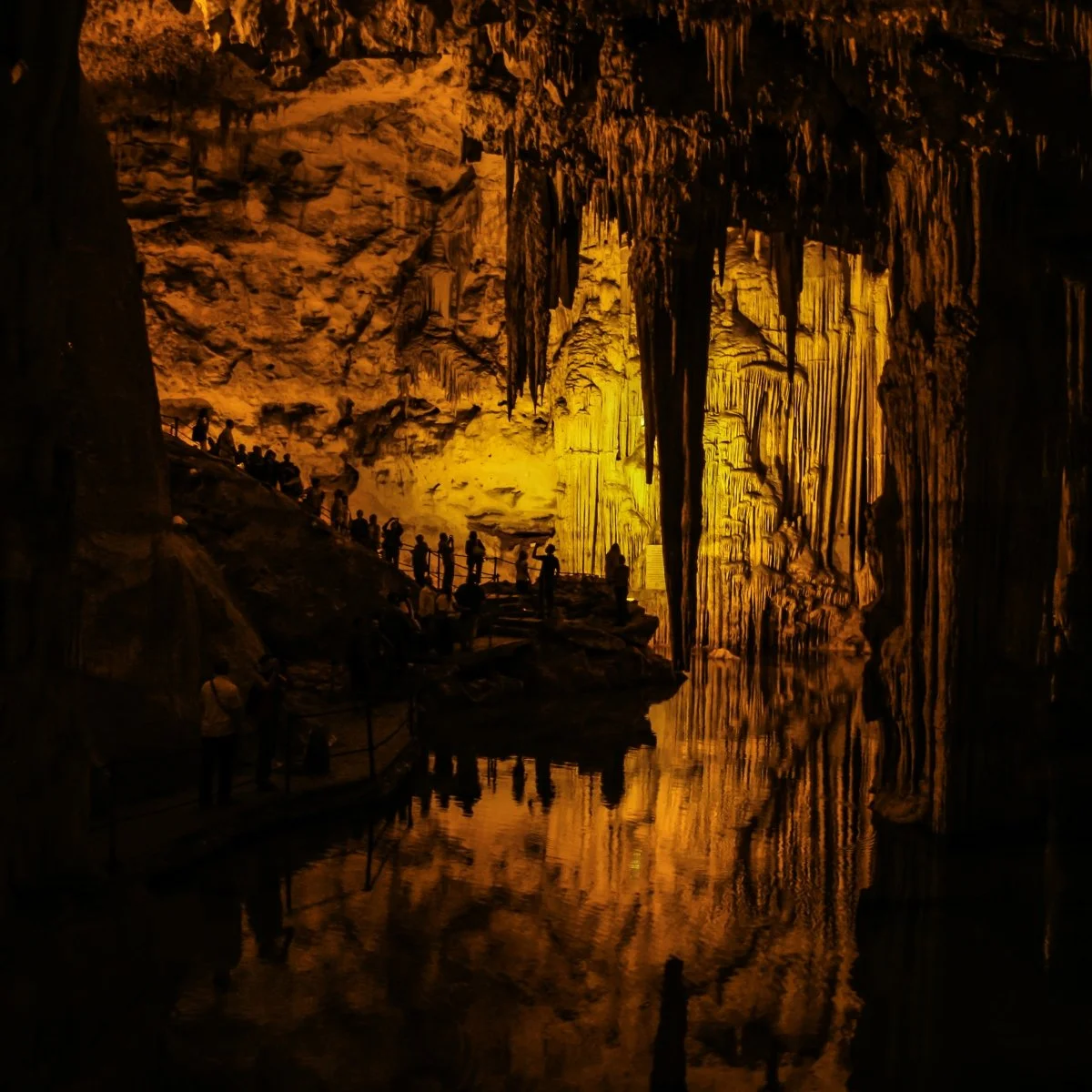 People walking inside a large illuminated cave with stalactites and stalagmites, and a reflective pool of water on the cave floor.