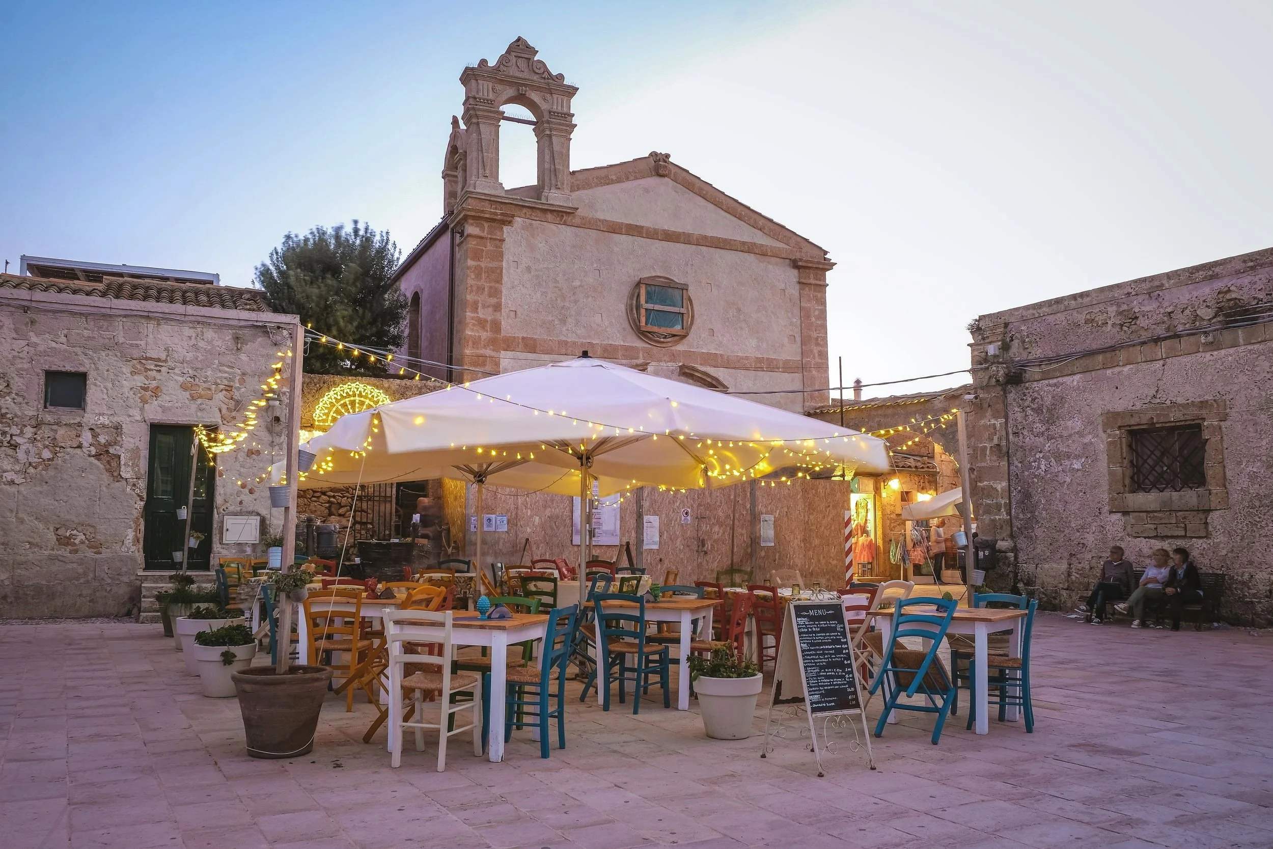 An outdoor dining area with multicolored chairs and white tables under a large white umbrella decorated with string lights in a historic stone town square, with a church or old building in background and people sitting on benches.