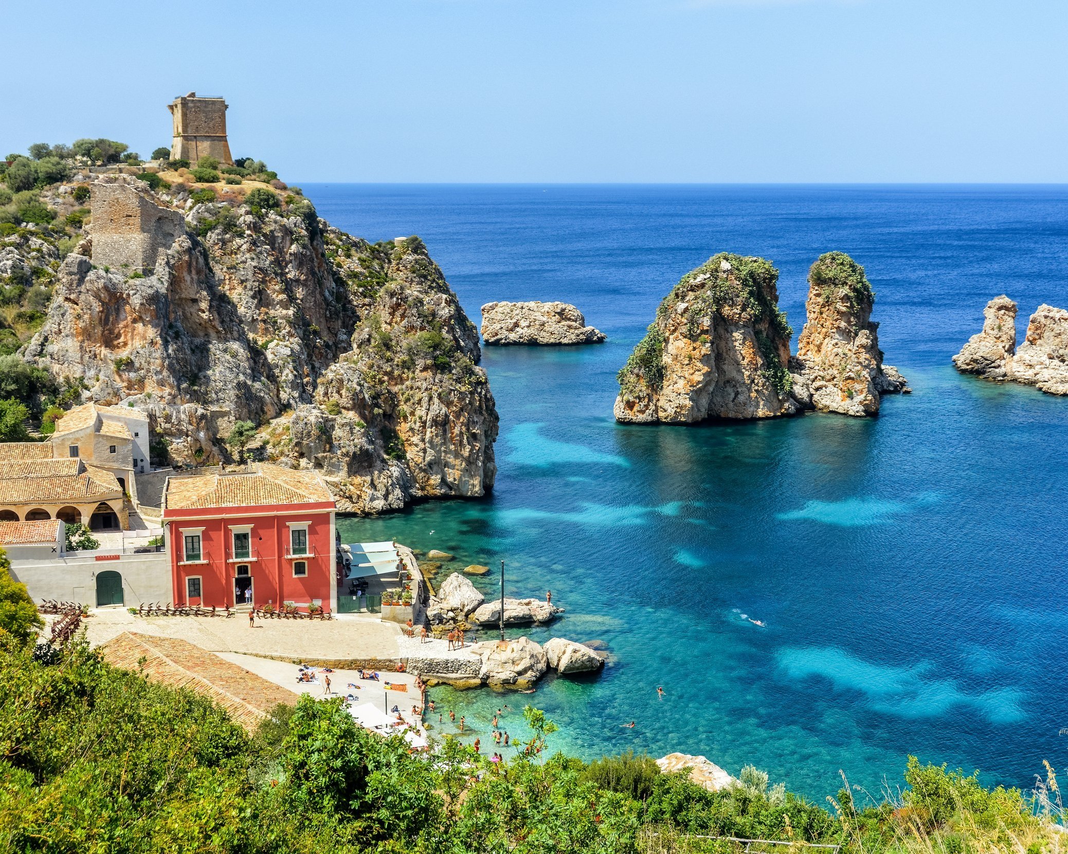 SCopello Coastal scene of a small village with colorful buildings beside turquoise water, rocky cliffs, and large sea stacks, under a clear blue sky.