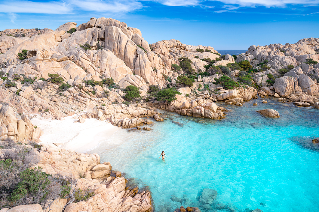 A woman swimming in a small, clear blue cove surrounded by rocky cliffs and sparse green shrubbery under a blue sky.