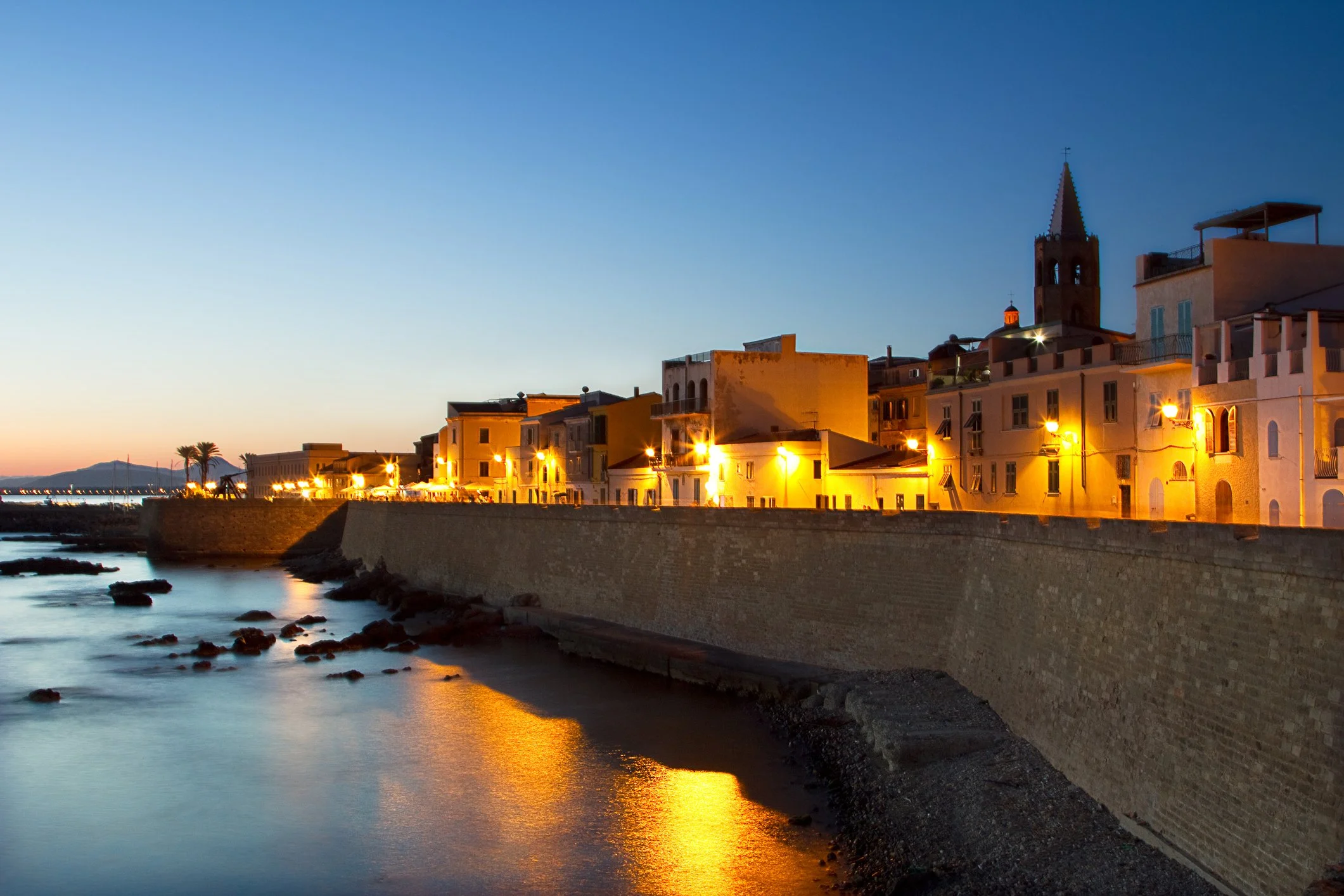 A seaside town at dusk with illuminated buildings along a waterfront, a brick seawall, calm water reflecting the streetlights, and a mountain in the distance.