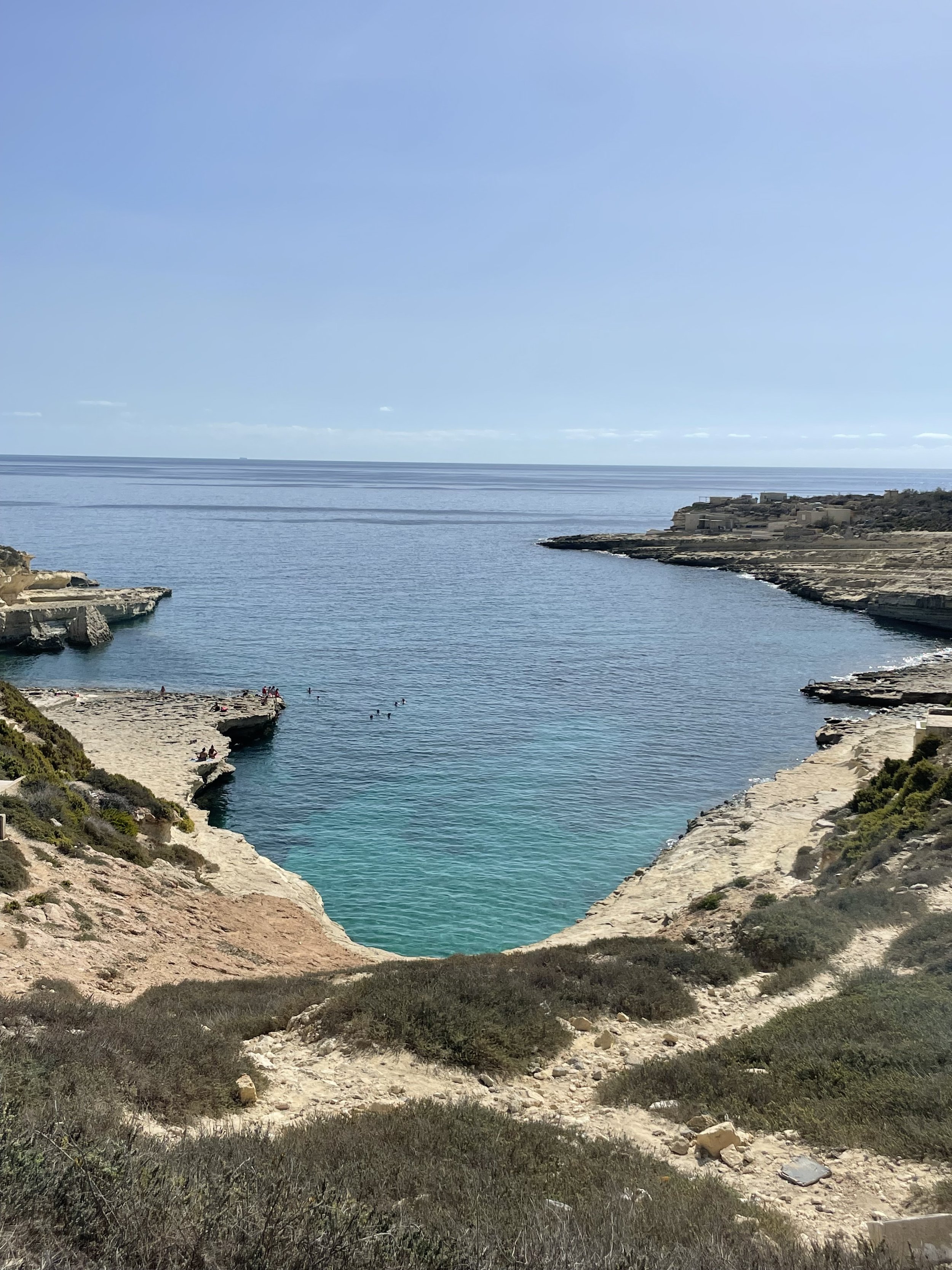 A secluded cove with clear turquoise water surrounded by rocky cliffs and sparse green vegetation, with a sandy beach on the left where people are relaxing, under a clear blue sky.