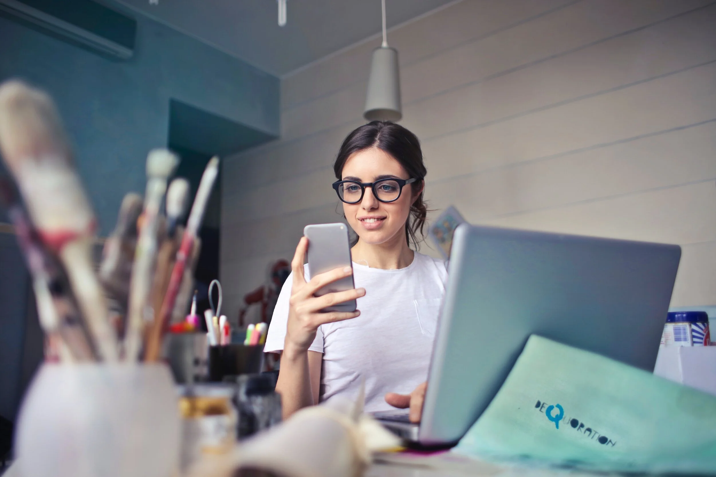 young woman wearing glasses with open laptop, looking at mobile phone, art supplies on desk