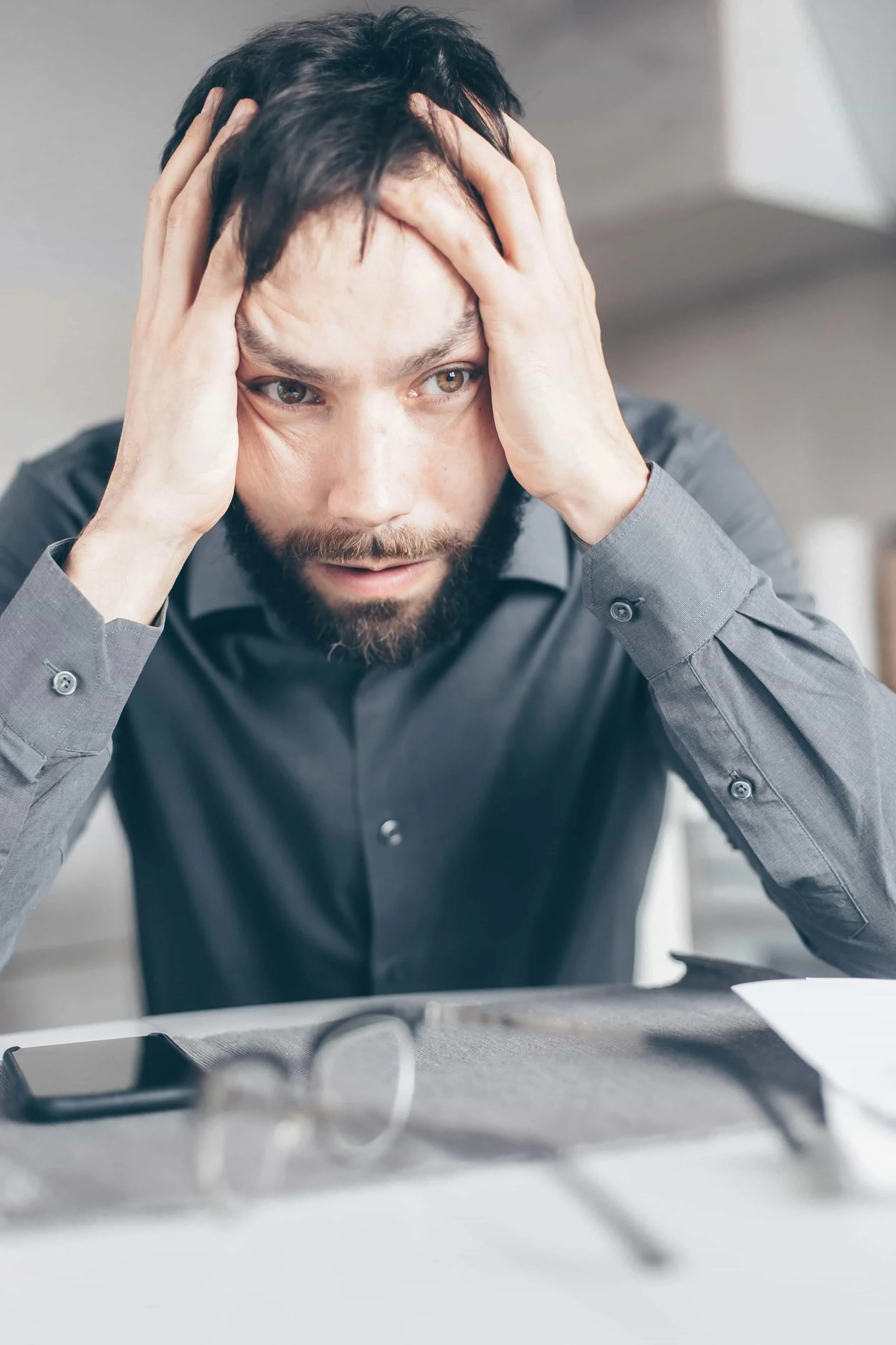 white man sitting at white table wearing dark grey shirt with head in his hands looking stressed