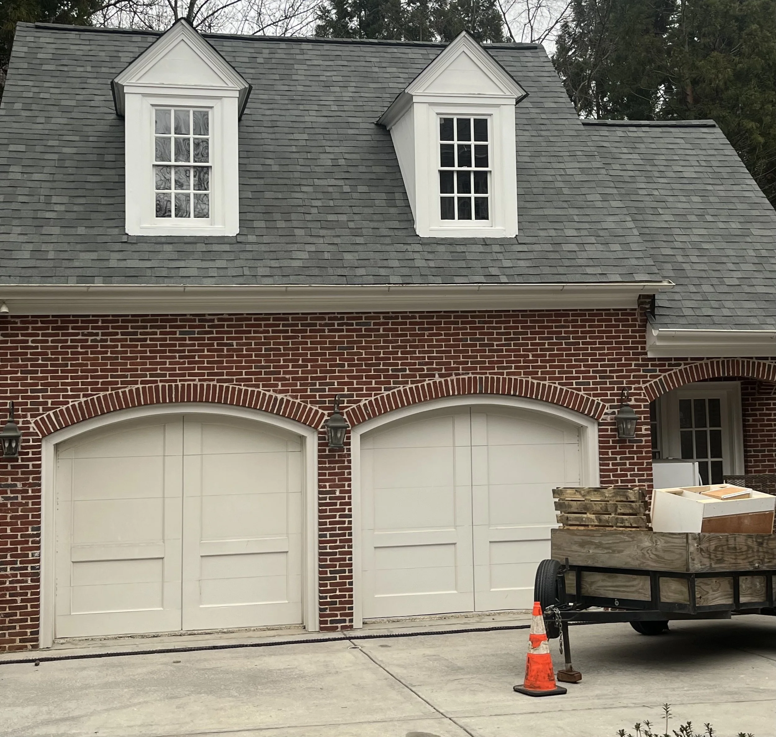 A house with a brick facade and two white garage doors, two dormer windows with white frames on the gray shingle roof, and a trailer with construction materials parked in front, alongside an orange traffic cone.
