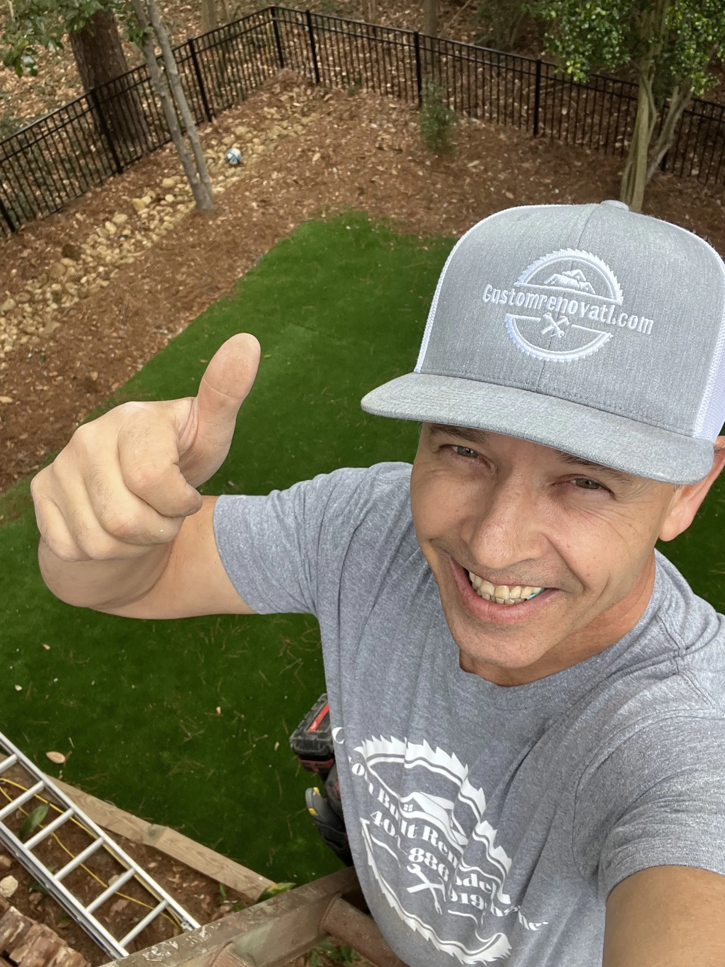 A man smiling and giving a thumbs up, wearing a gray cap and gray t-shirt, standing on a ladder in a backyard with a fenced garden area and green grass.