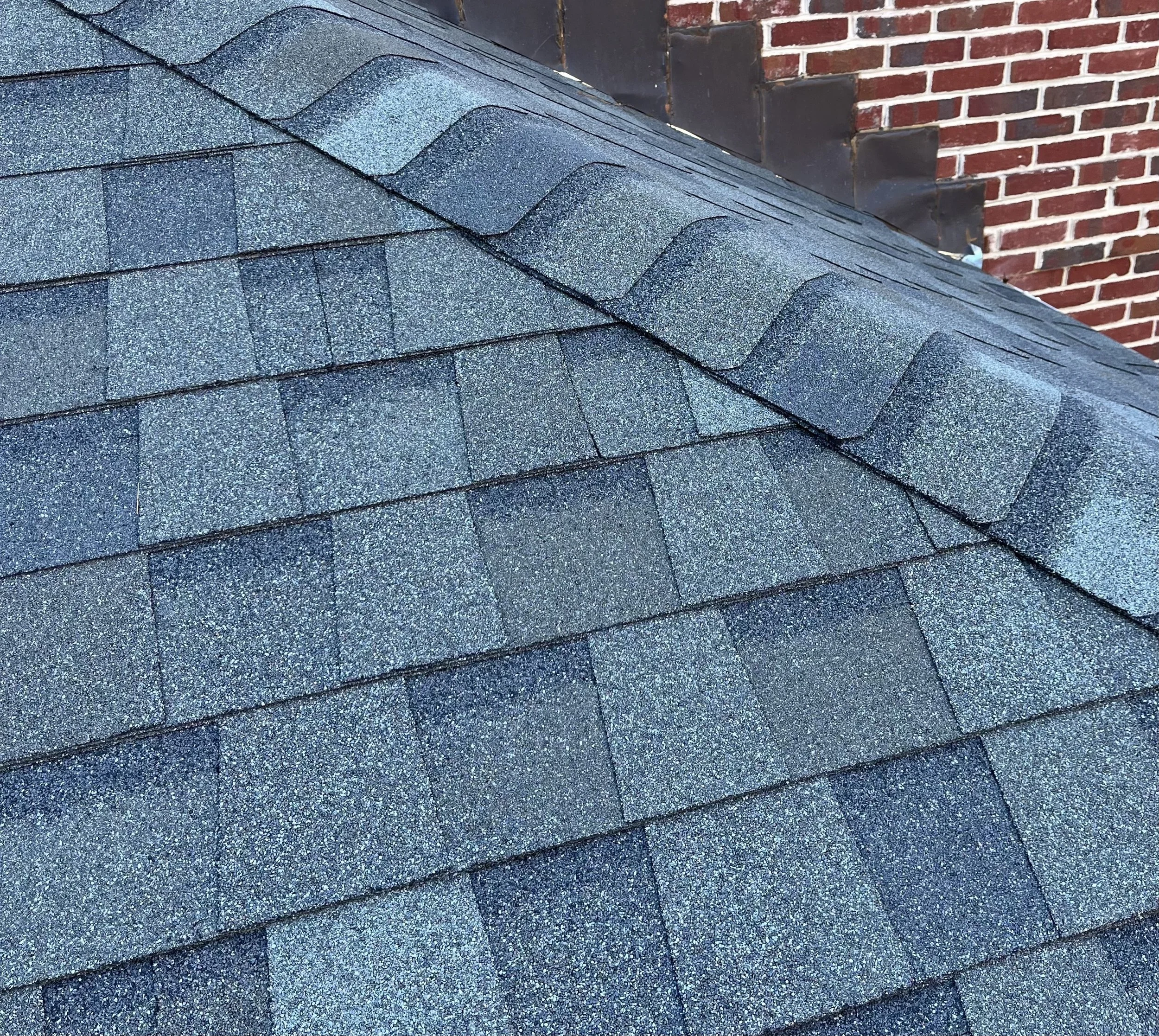 Close-up view of a shingle roof with greenish-black asphalt shingles and a brick chimney in the background.