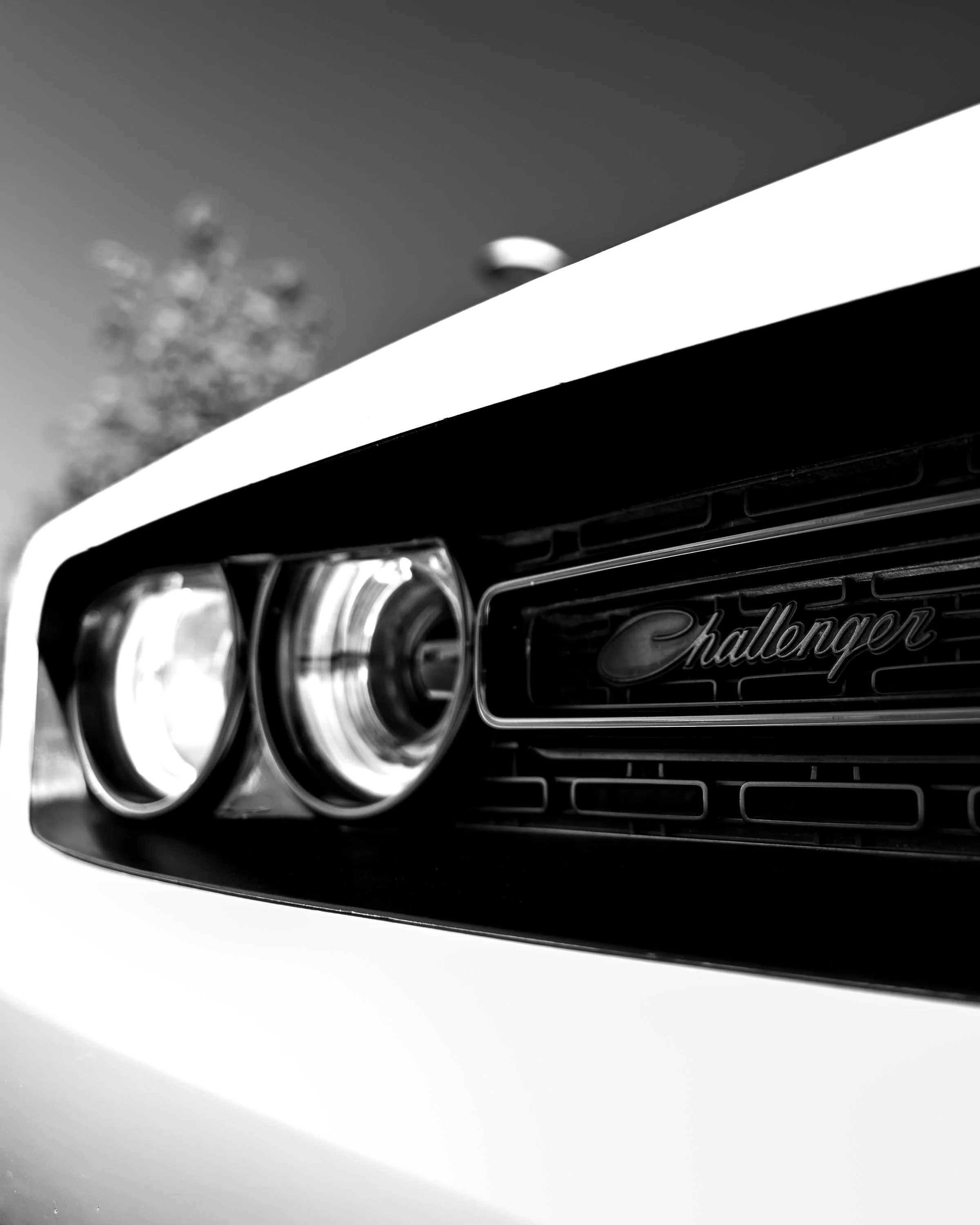 Close-up of a Dodge Challenger front grille with the brand's emblem, headlight, and part of the car's hood, taken in black and white.