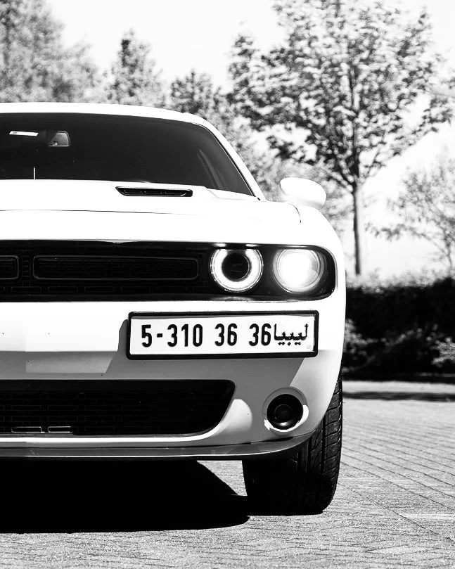 Close-up of the front of a white Dodge Challenger car with an Arabic license plate, parked outdoors with trees in the background.
