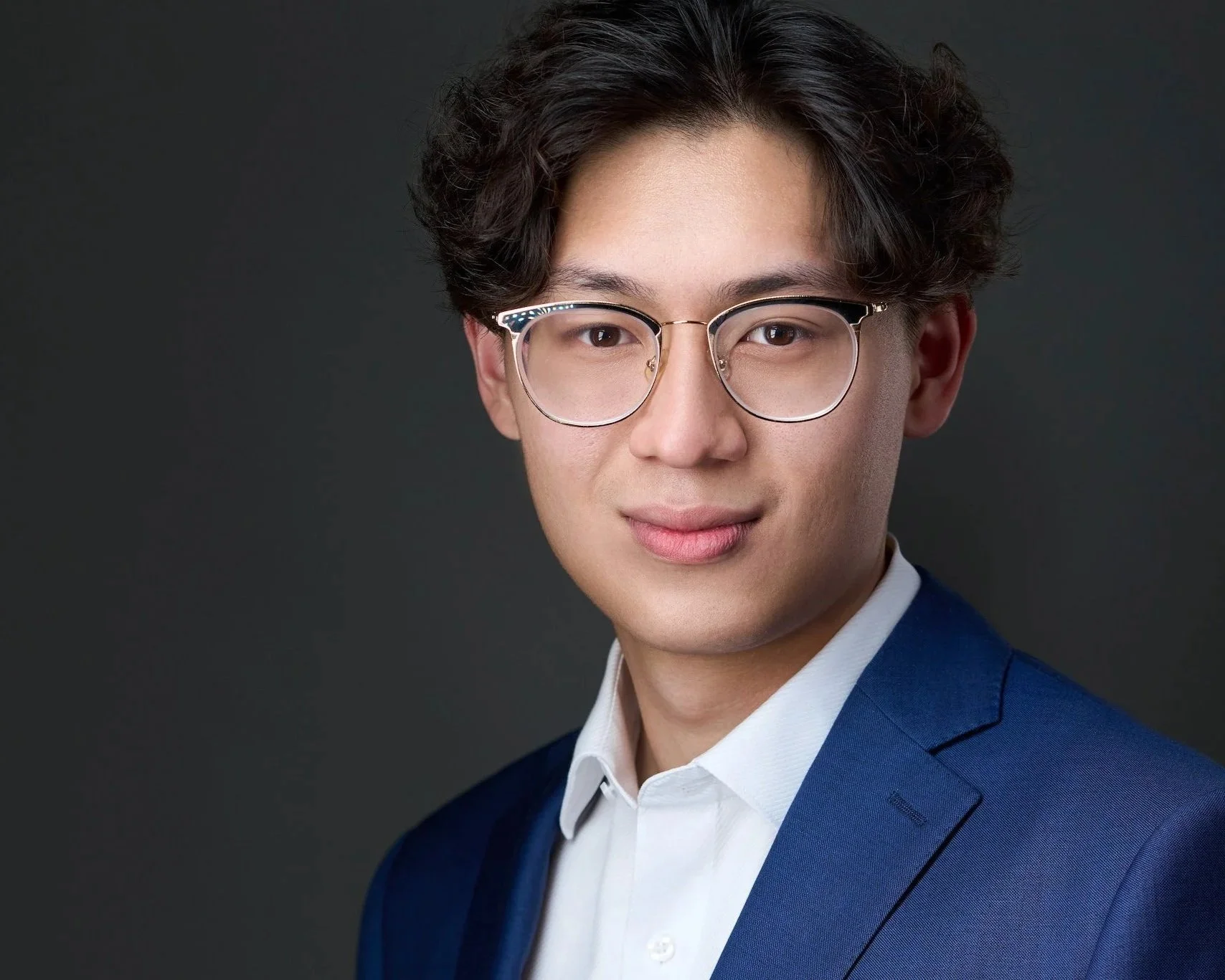 Portrait of a young man wearing glasses, a white shirt, and a blue blazer against a dark background.