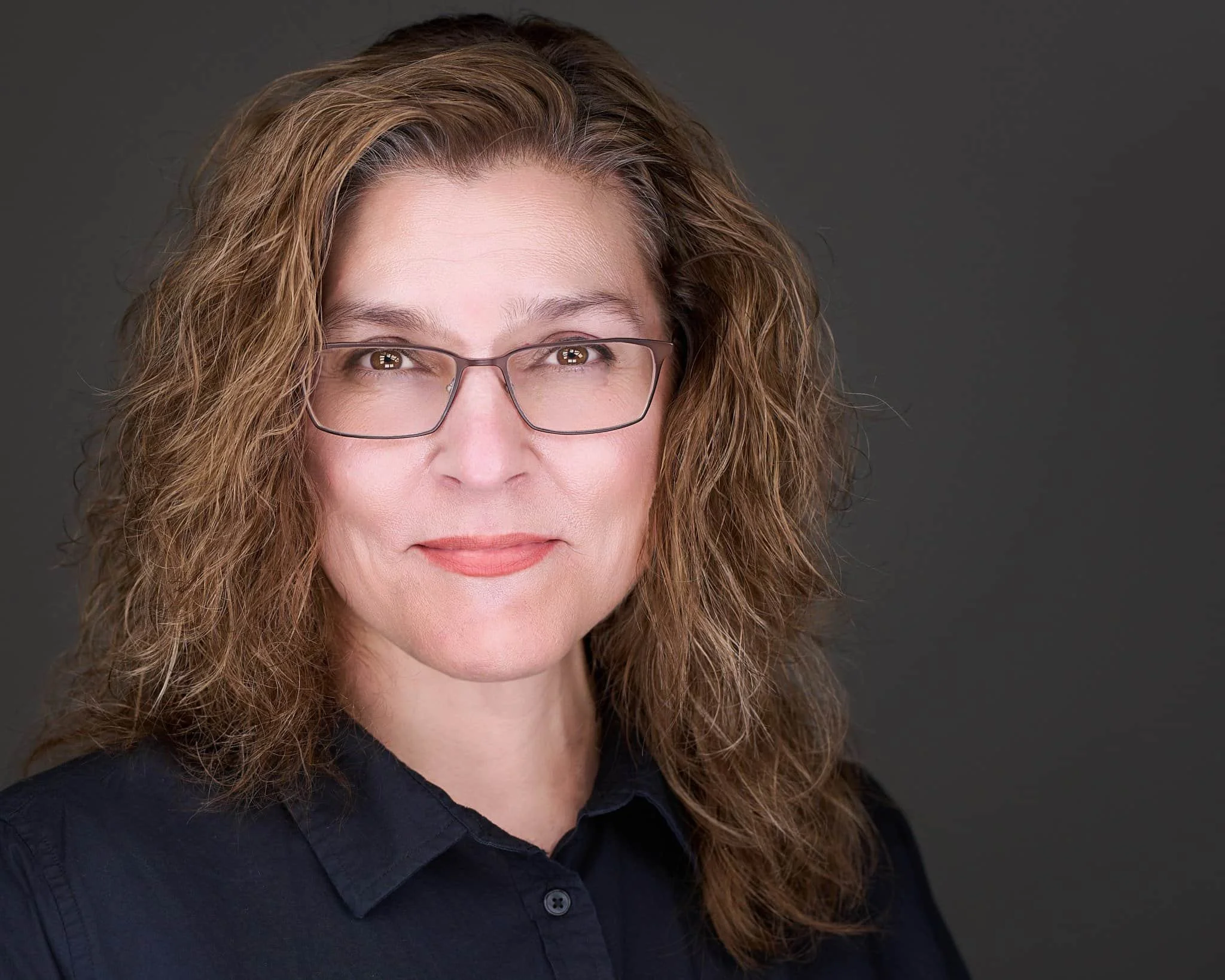 A woman with shoulder-length curly brown hair and glasses, wearing a black collared shirt, smiling softly against a dark gray background.