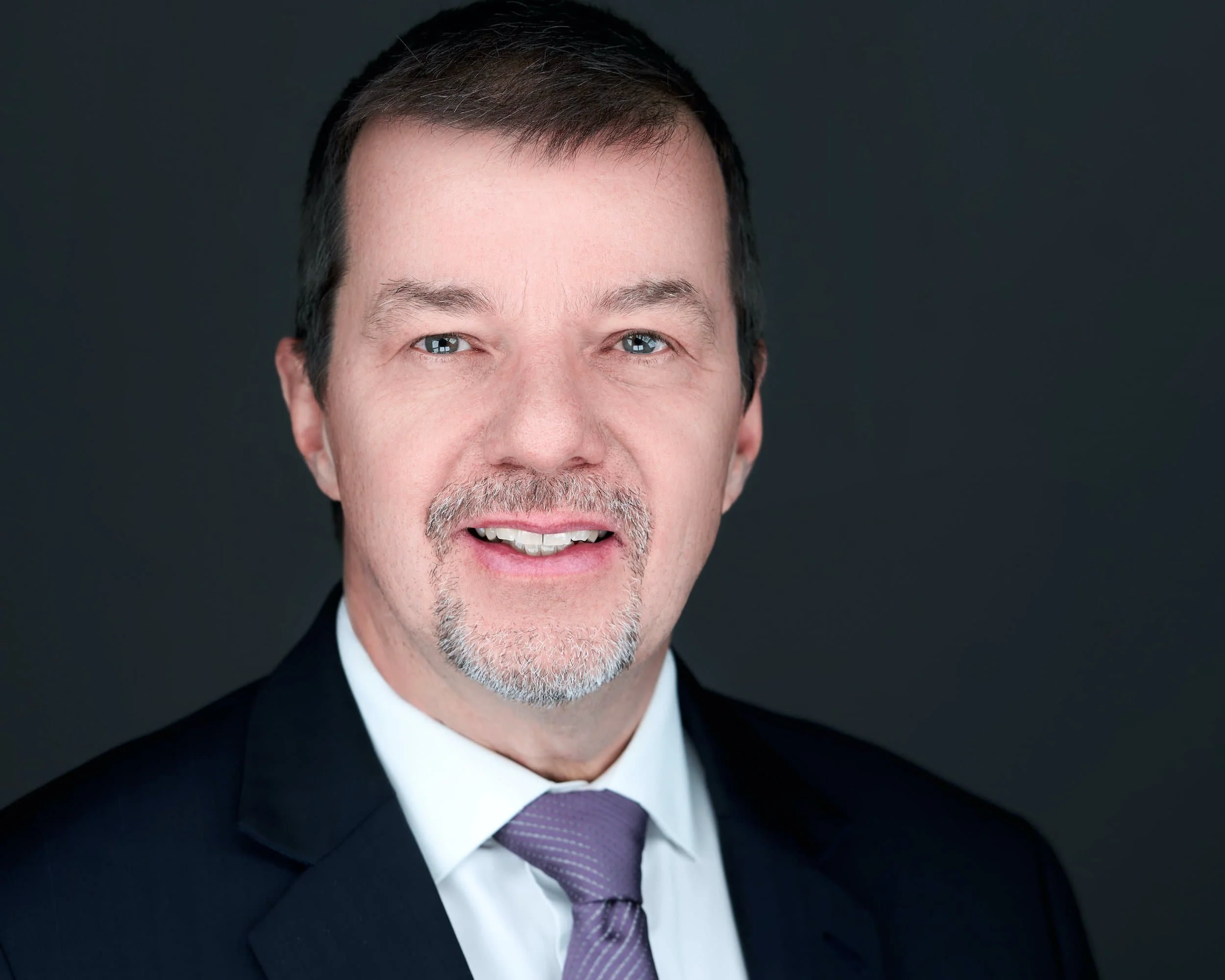 Headshot of a middle-aged man with short dark hair, gray beard, wearing a dark suit, white shirt, and purple tie, against a dark background.
