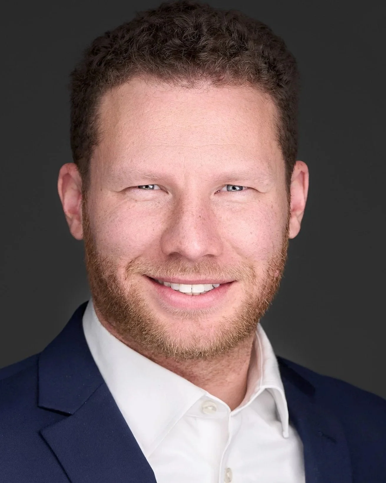 Headshot of a man with curly red hair, a beard, wearing a white dress shirt and a navy blazer against a dark background.