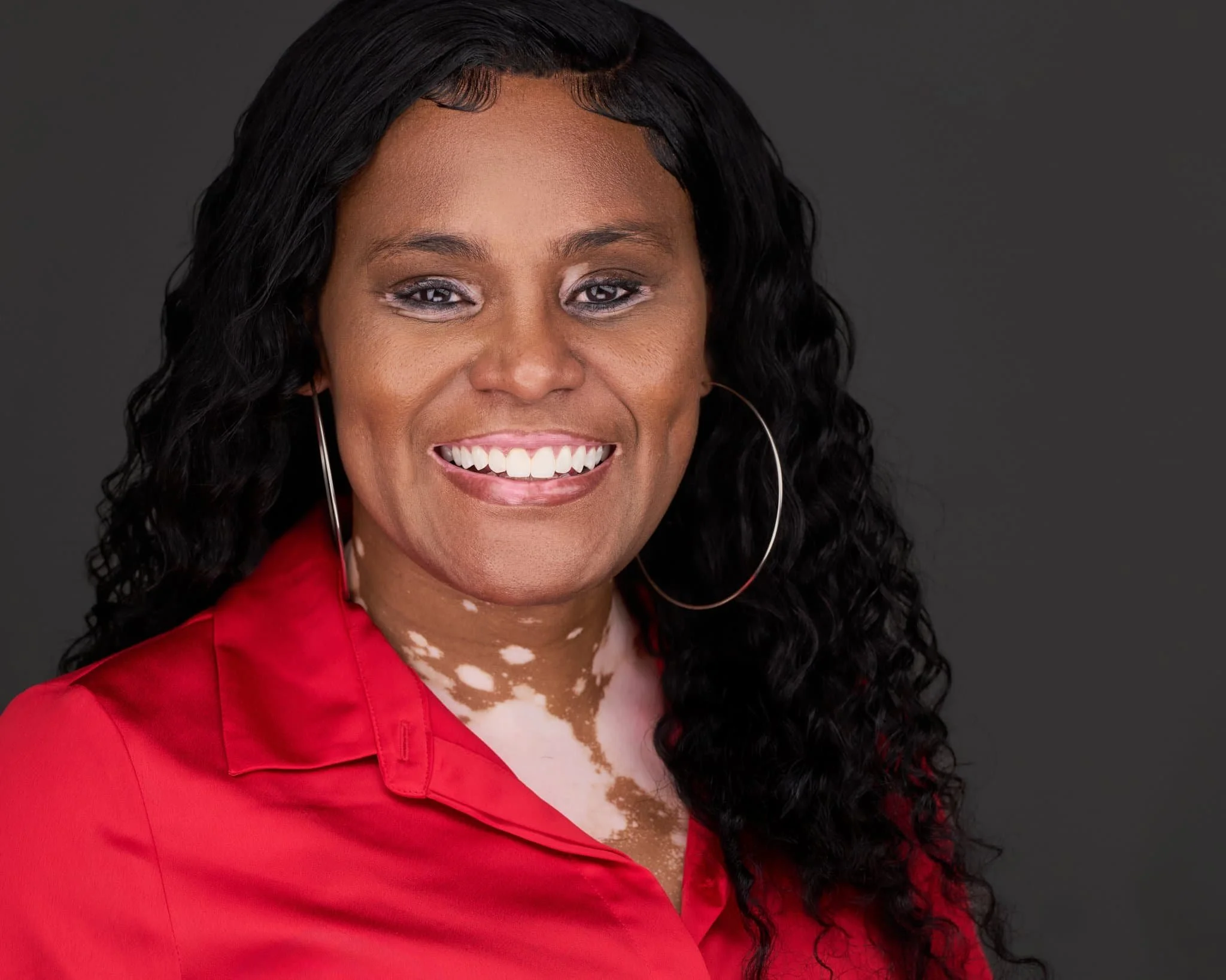 Close-up portrait of a smiling woman with curly black hair, wearing a red blouse and large hoop earrings, against a dark gray background.