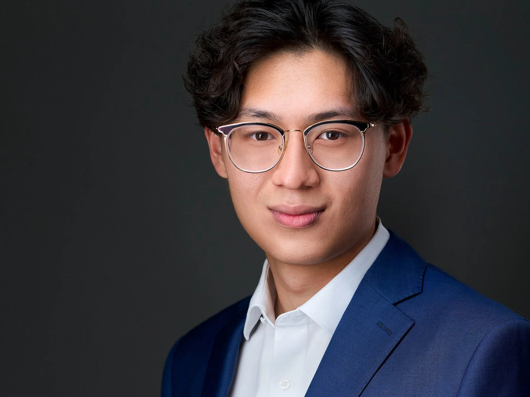 A young man dressed in a formal blue suit, white dress shirt, wearing round glasses, and looking at the camera with a slight smile, against a dark background.