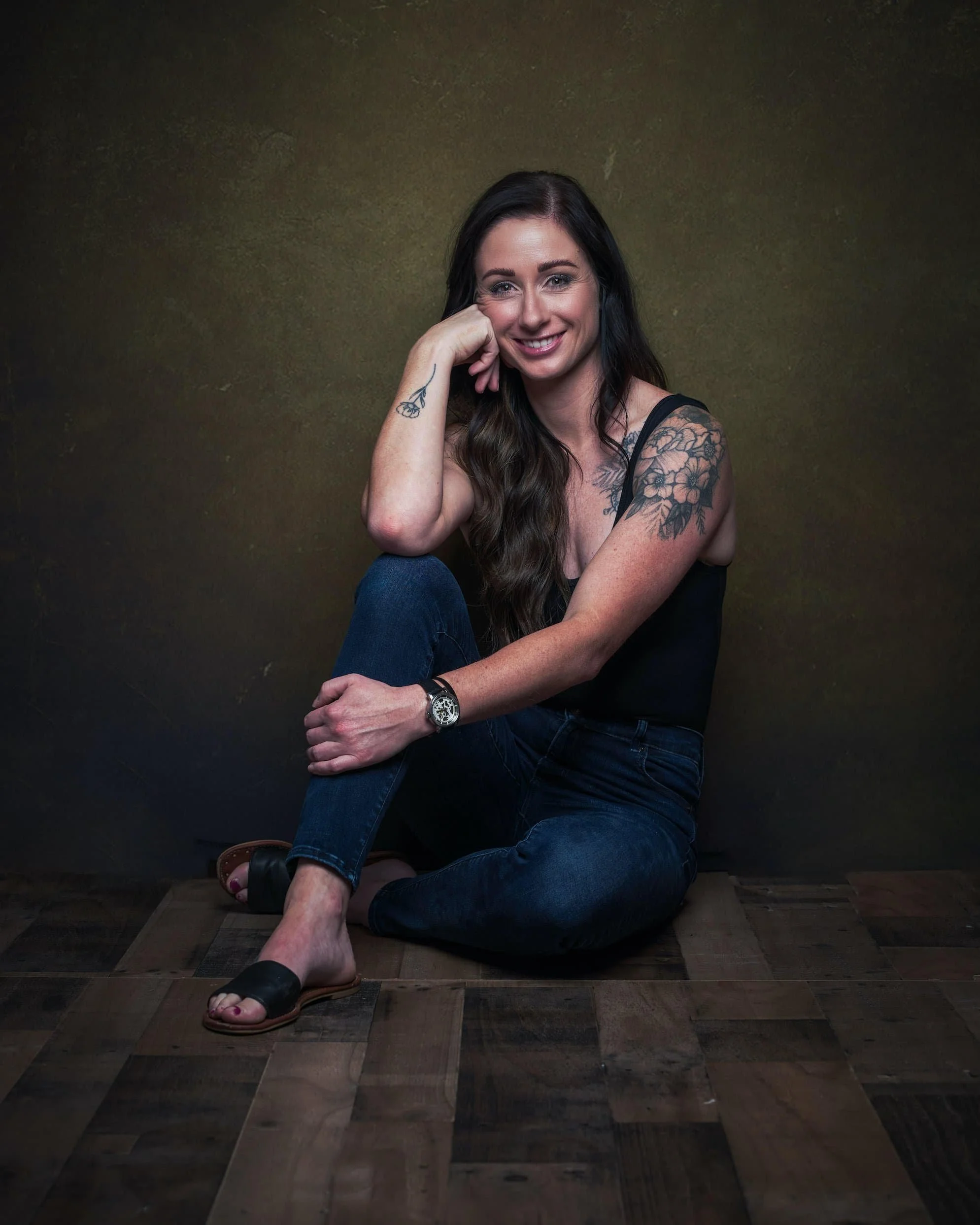A woman with long dark hair and tattoos, sitting on a wooden floor against a dark background, smiling at the camera. She is wearing a black tank top, blue jeans, and sandals.