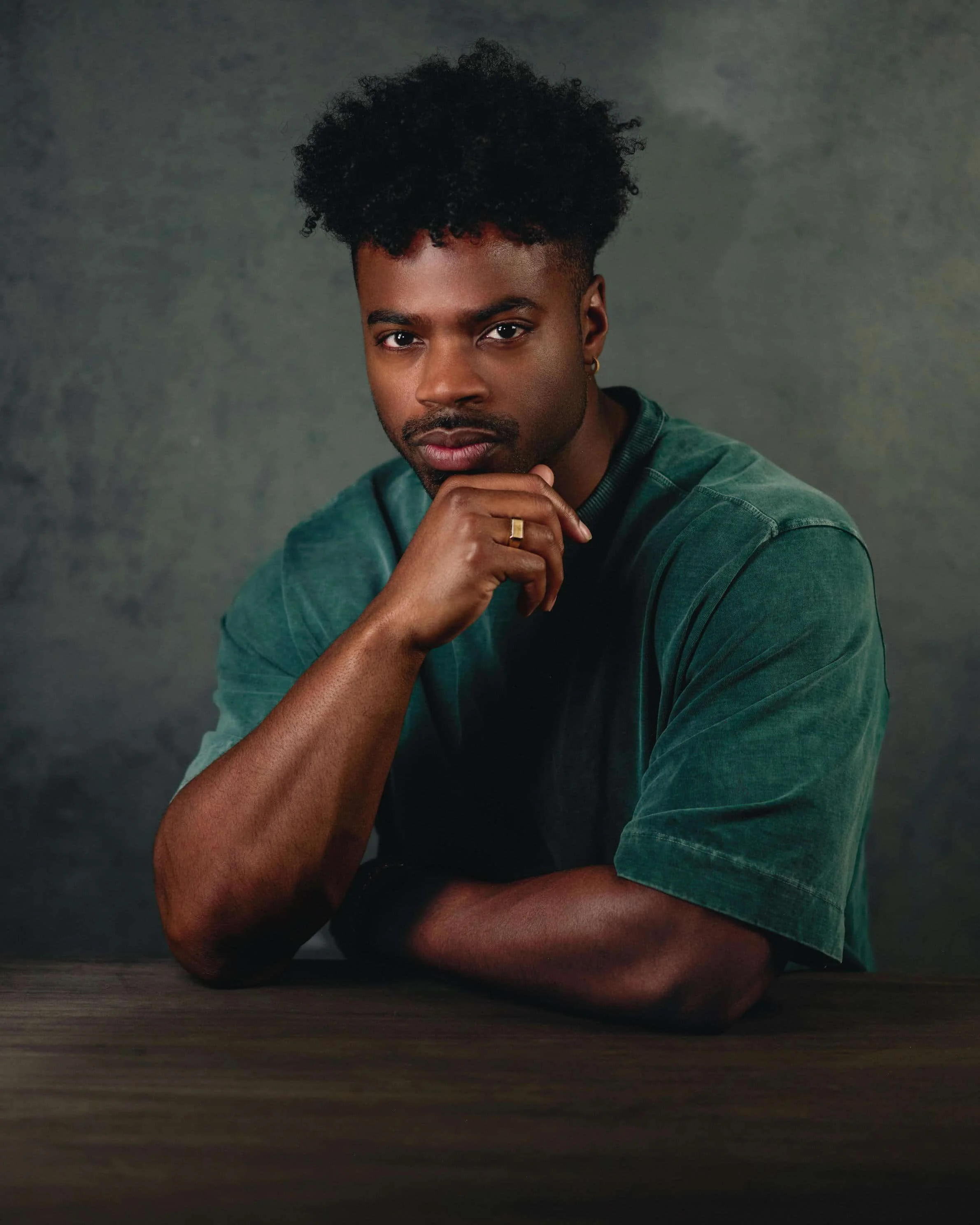 A young man with curly black hair and a short beard, wearing a green T-shirt, sitting at a wooden table, resting his chin on his hand, looking directly at the camera.