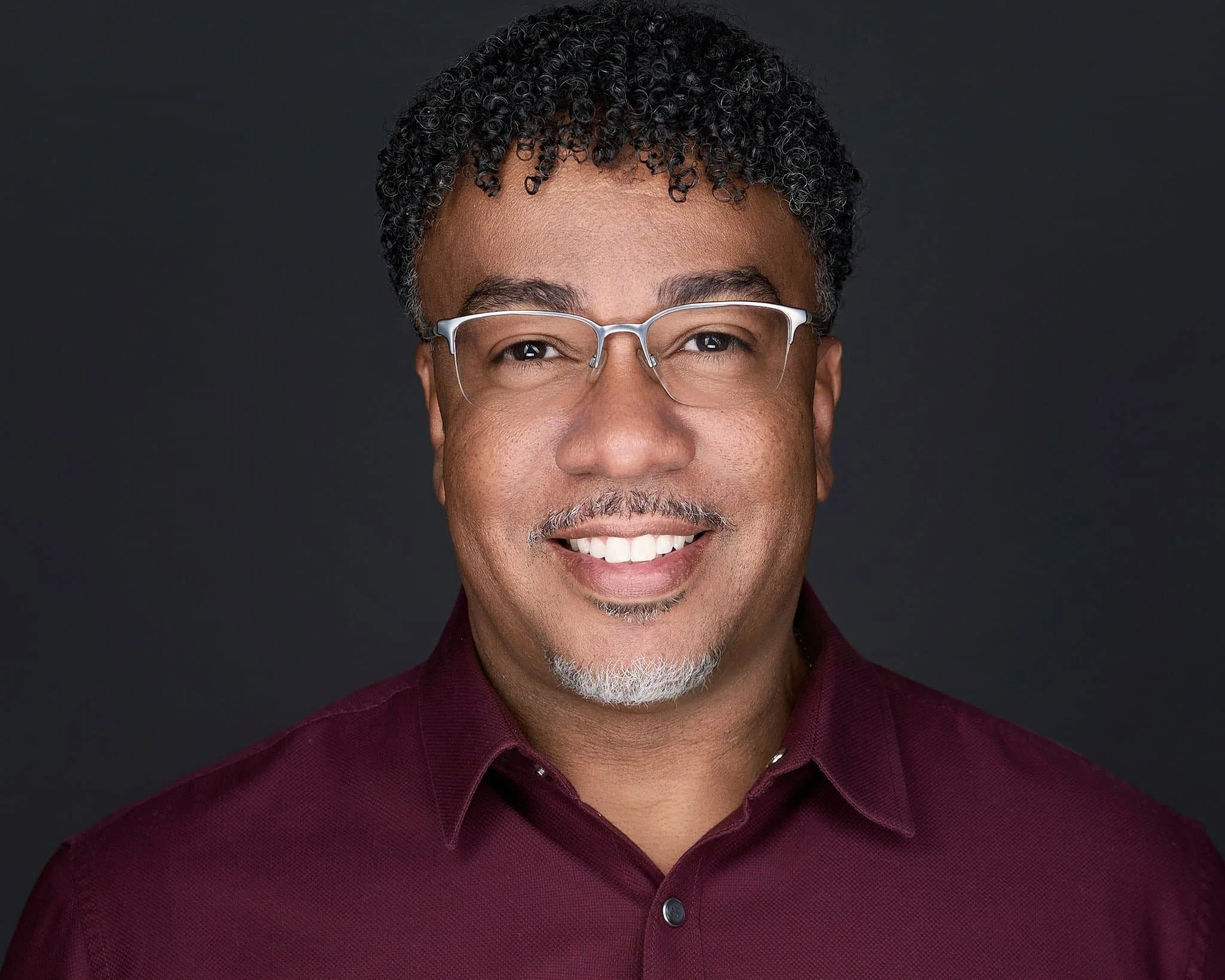 A middle-aged man with curly black and gray hair, wearing clear eyeglasses and a maroon collared shirt, smiling against a dark background.