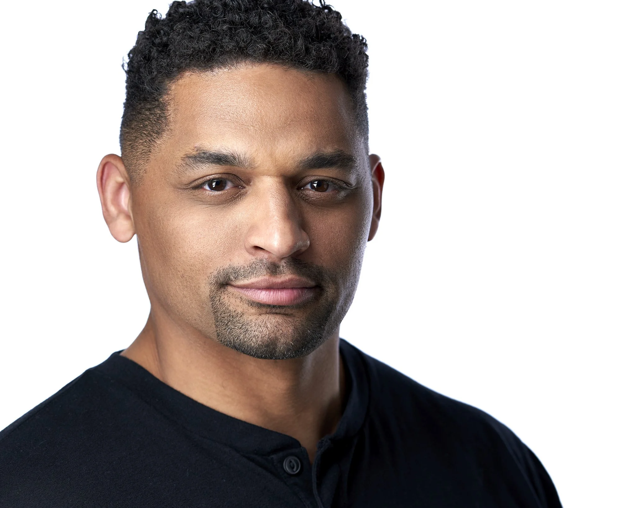 Close-up portrait of a man with short, curly black hair, brown skin, and wearing a black shirt, smiling subtly with a white background.