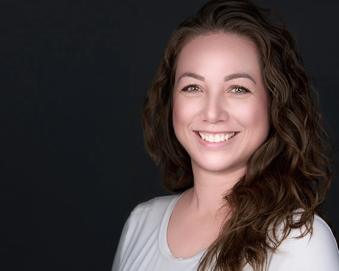 A smiling woman with curly brown hair wearing a white top against a dark background.