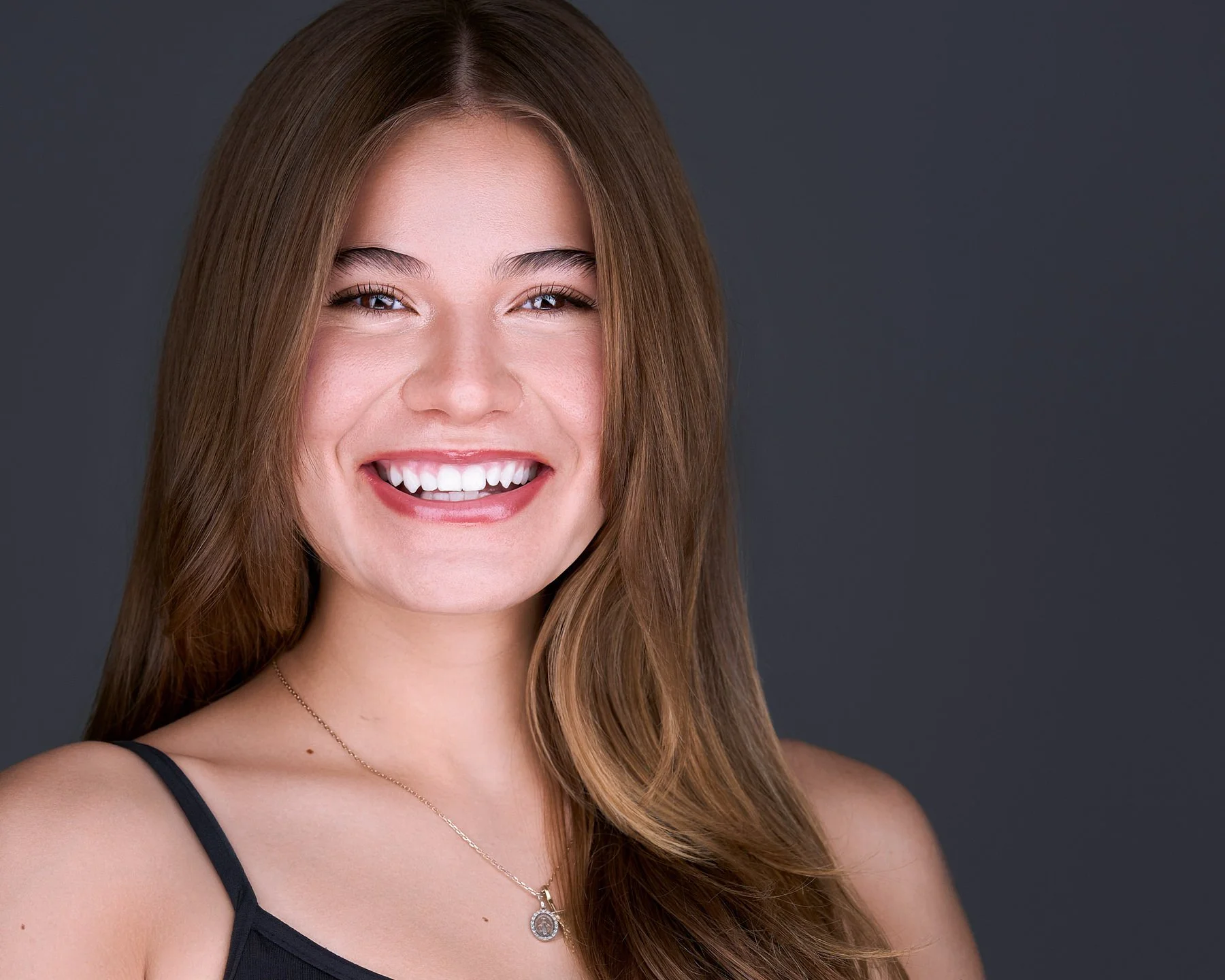 A young woman with long, wavy brown hair smiling against a dark gray background, wearing a black spaghetti strap top and a silver necklace with a pendant.