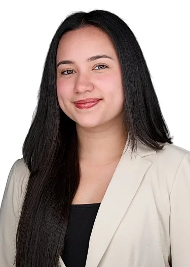 Portrait of a young woman with long black hair, wearing a light-colored blazer over a black top, smiling at the camera.