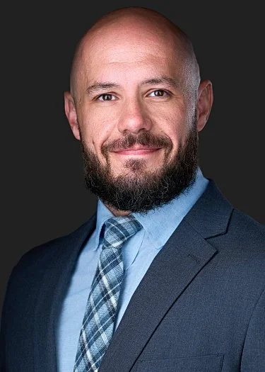 Professional portrait of a bald man with a beard, wearing a dark suit and light blue shirt with a plaid tie, against a dark background.