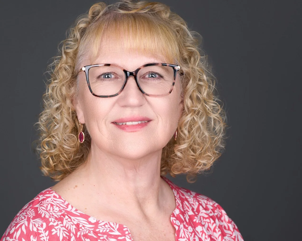 A middle-aged woman with curly blonde hair, wearing glasses, earrings, and a pink floral top, smiling against a dark grey background.