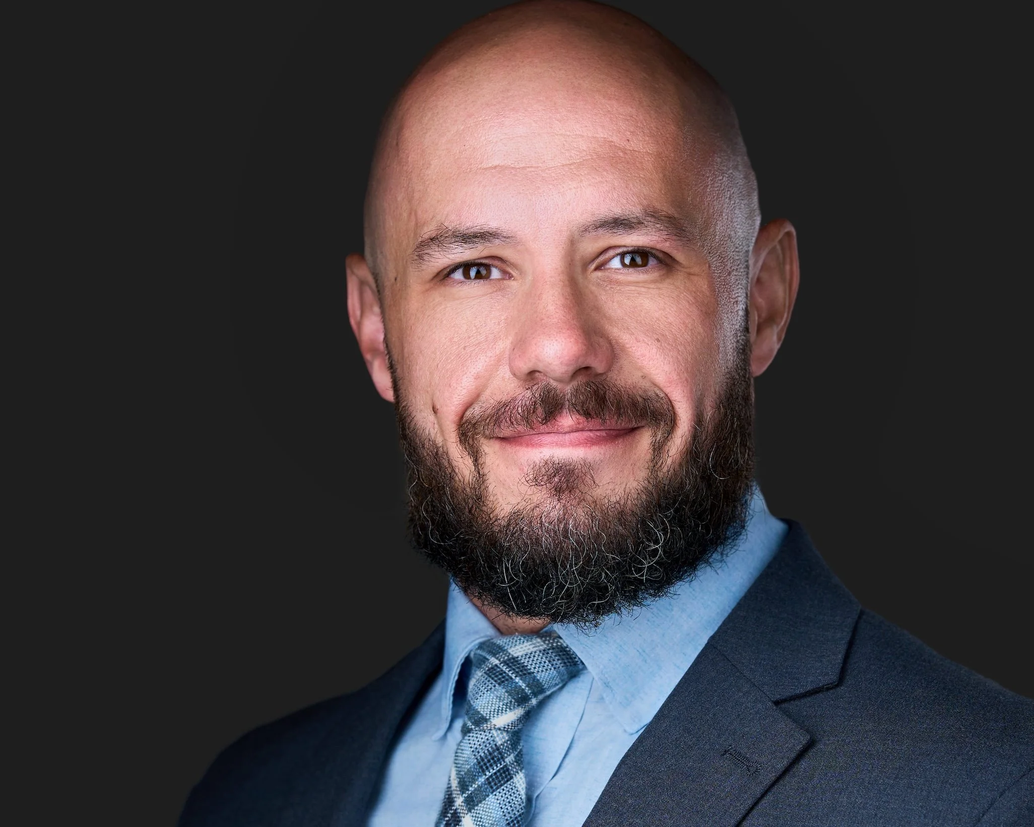 Close-up portrait of a man with a bald head and a well-groomed beard dressed in a dark suit, light blue shirt, and a checkered tie, smiling slightly against a dark background.