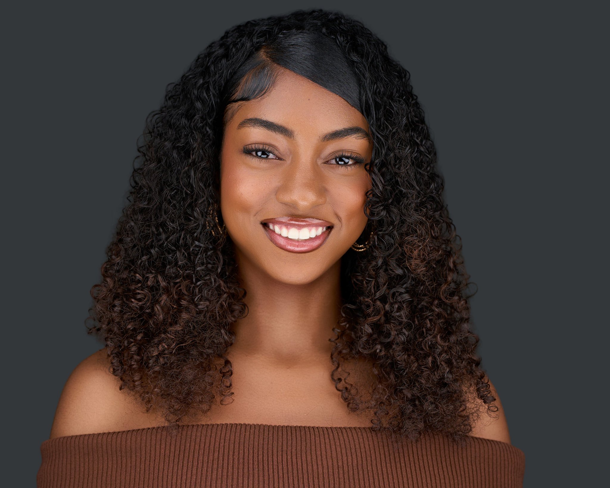 Portrait of a young woman smiling with long curly black hair, wearing a brown top against a dark gray background.