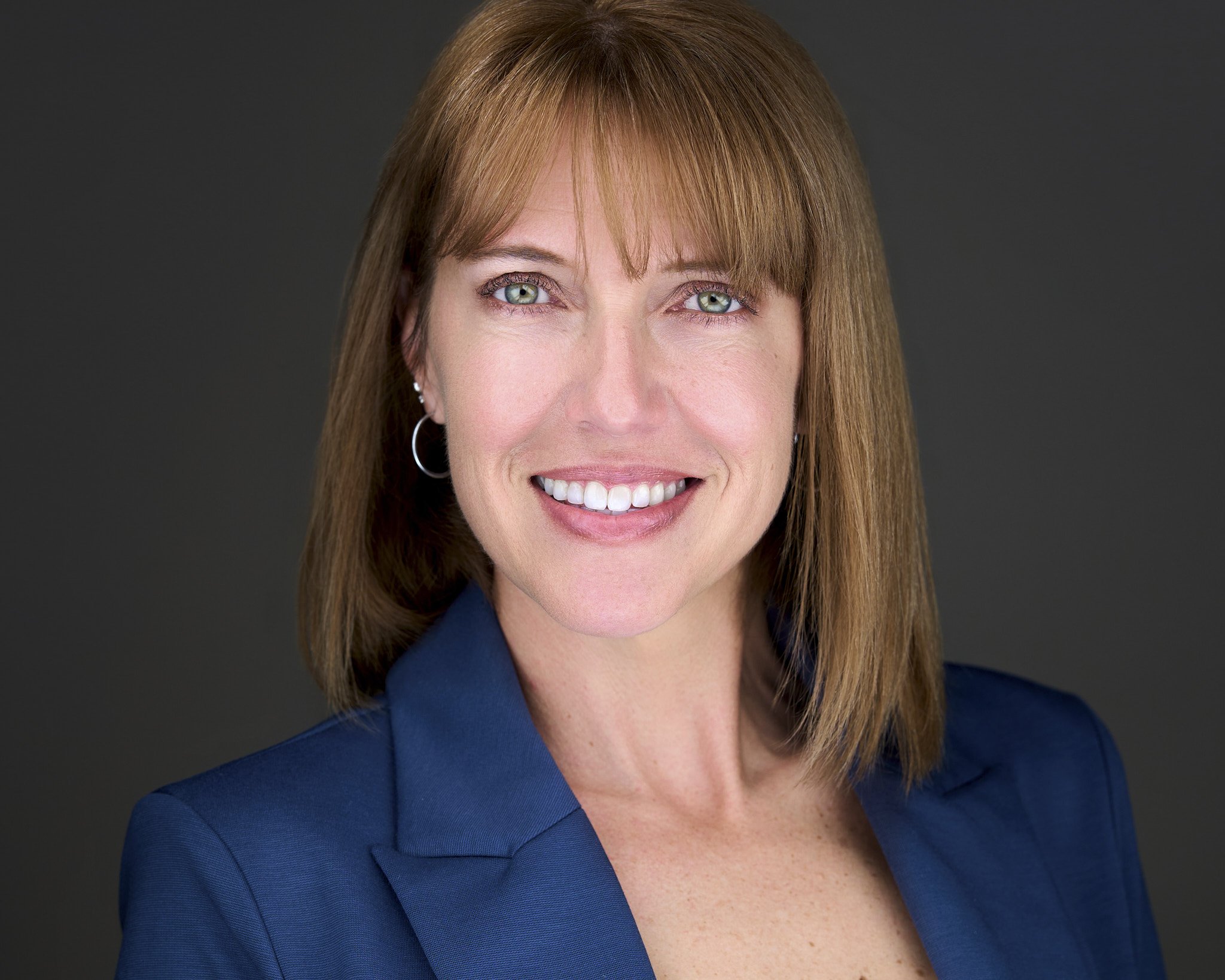 Close-up of a woman with shoulder-length red hair, wearing a blue blazer, smiling, with earrings, against a gray background.