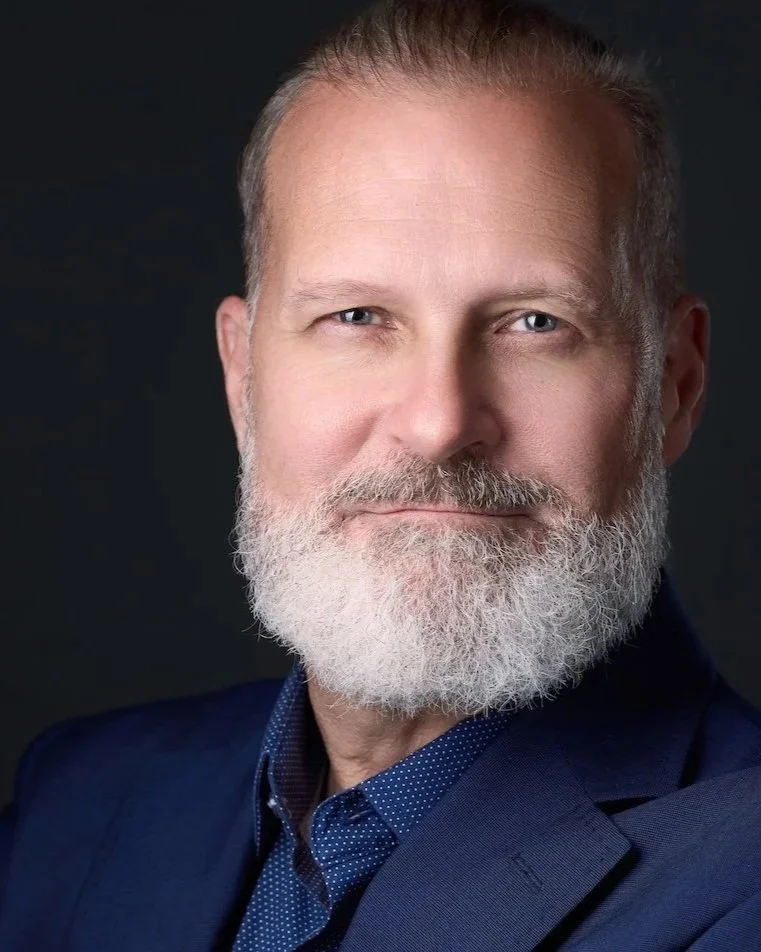 Close-up portrait of a middle-aged man with a full gray beard and short gray hair, wearing a dark navy suit and a blue polka dot shirt, against a black background.