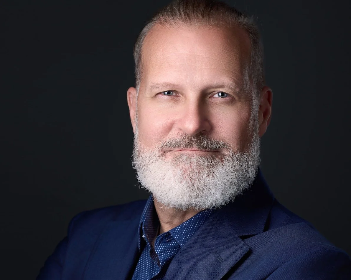 Close-up portrait of a middle-aged man with a gray beard and short hair, wearing a dark blue suit and a patterned dress shirt, posing against a dark background.