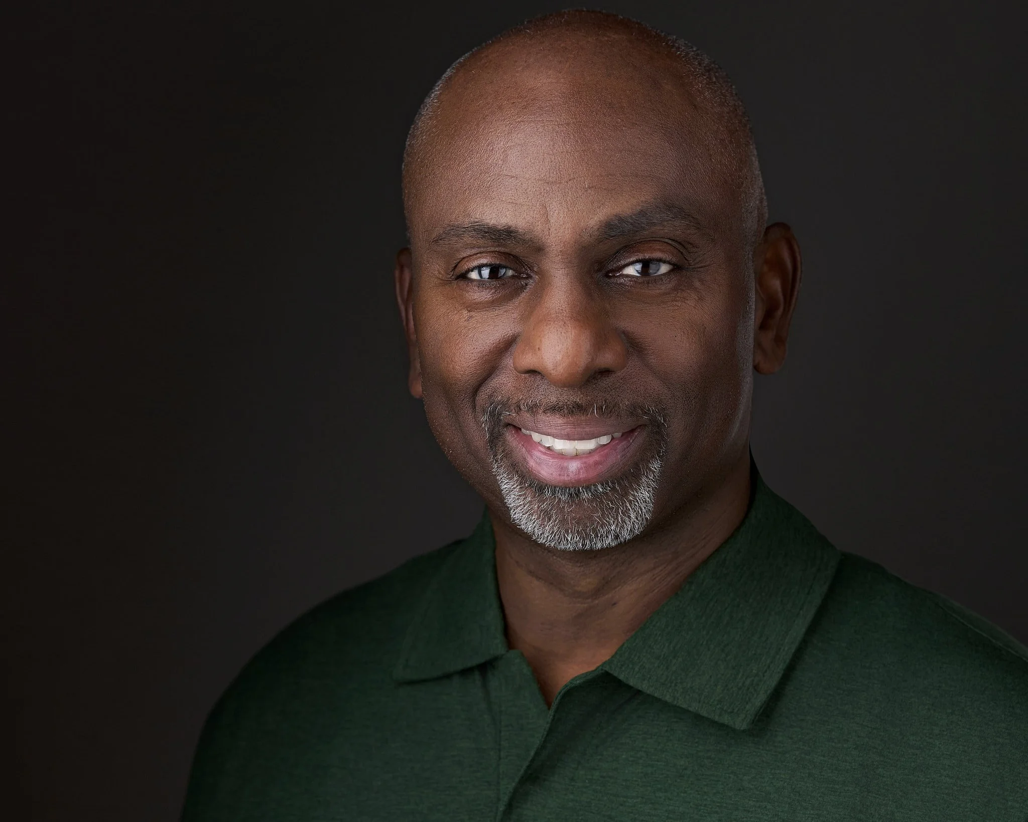 Close-up of a smiling middle-aged African American man with blue eyes, short gray hair, and a gray beard, wearing a dark green sweater, against a black background.