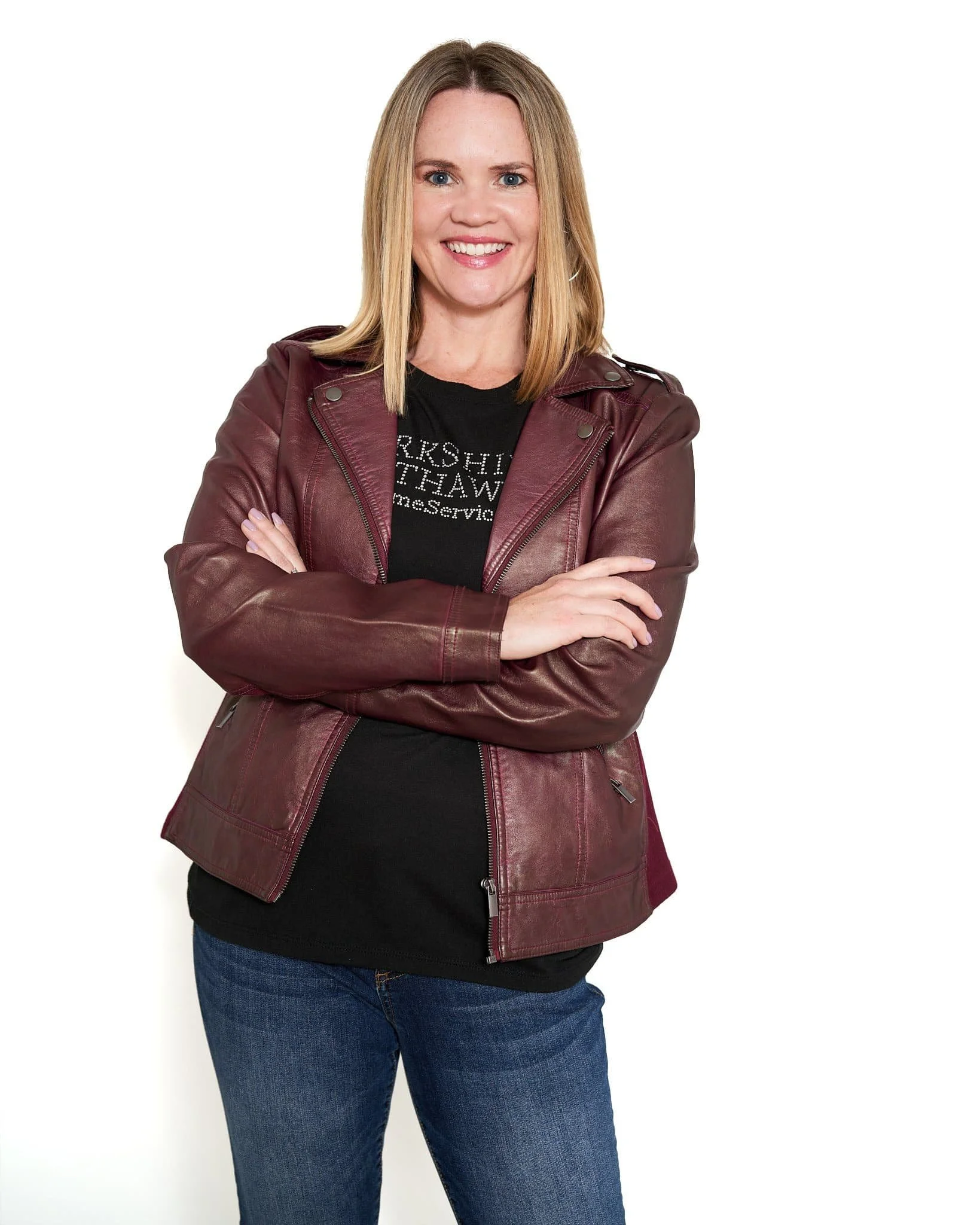 A woman with shoulder-length blonde hair smiling, arms crossed, wearing a maroon leather jacket over a black t-shirt and blue jeans, standing against a white background.