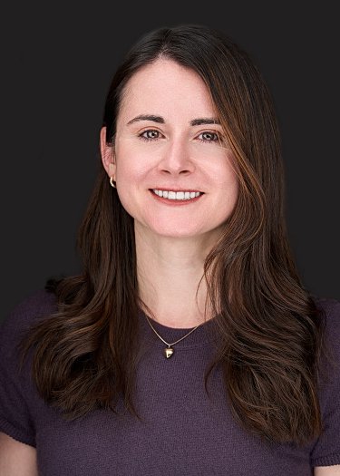 Portrait of a woman with long brown hair, wearing a dark purple top and a necklace, smiling against a dark background.