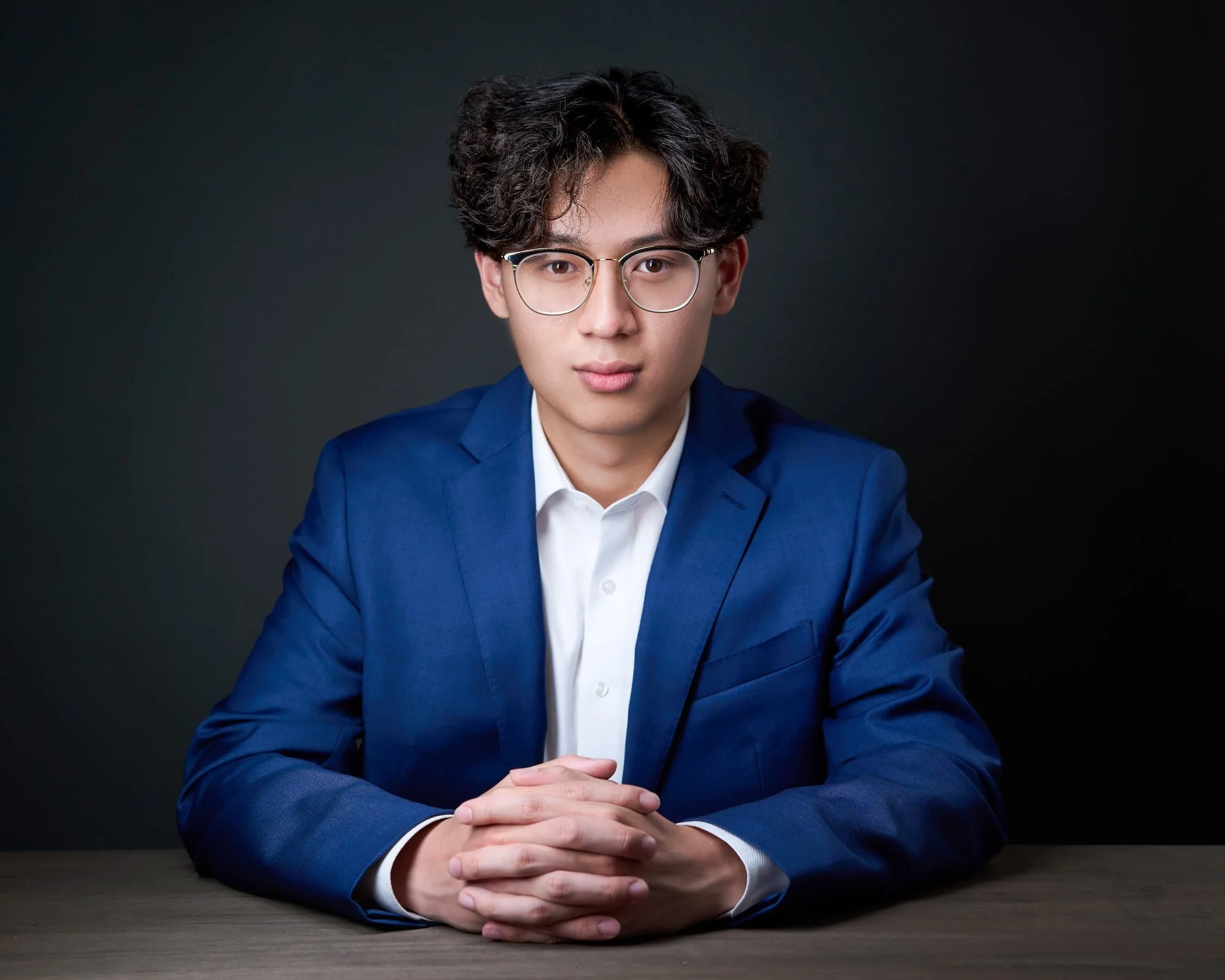 Young man with dark curly hair, glasses, dressed in a blue suit and white shirt, sitting at a table with hands clasped, against a dark background.