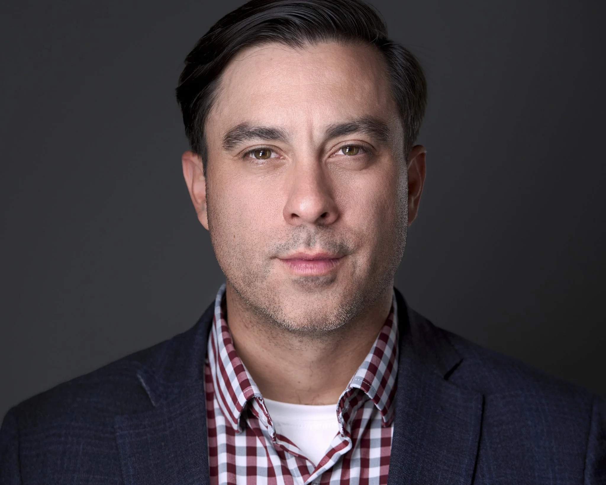 Close-up portrait of a young man with short dark hair, hazel eyes, wearing a checkered shirt and dark cardigan, against a dark background.