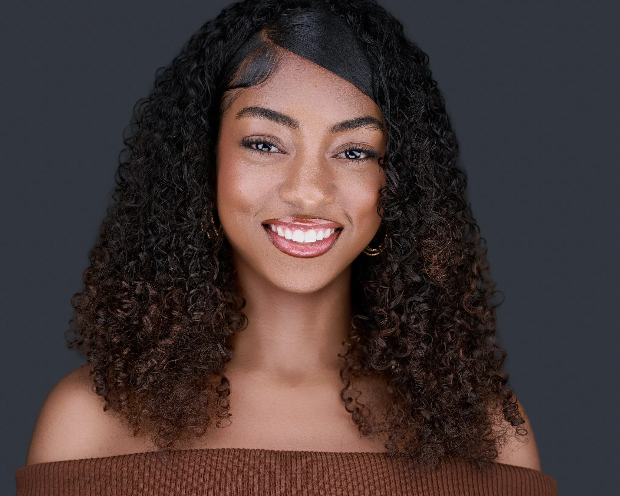 A young woman with curly black hair and a silky black wig at the front, smiling broadly against a gray background. She has well-defined eyebrows, brown eyes, and is wearing light makeup and earrings. She is dressed in a sleeveless, brown ribbed top.