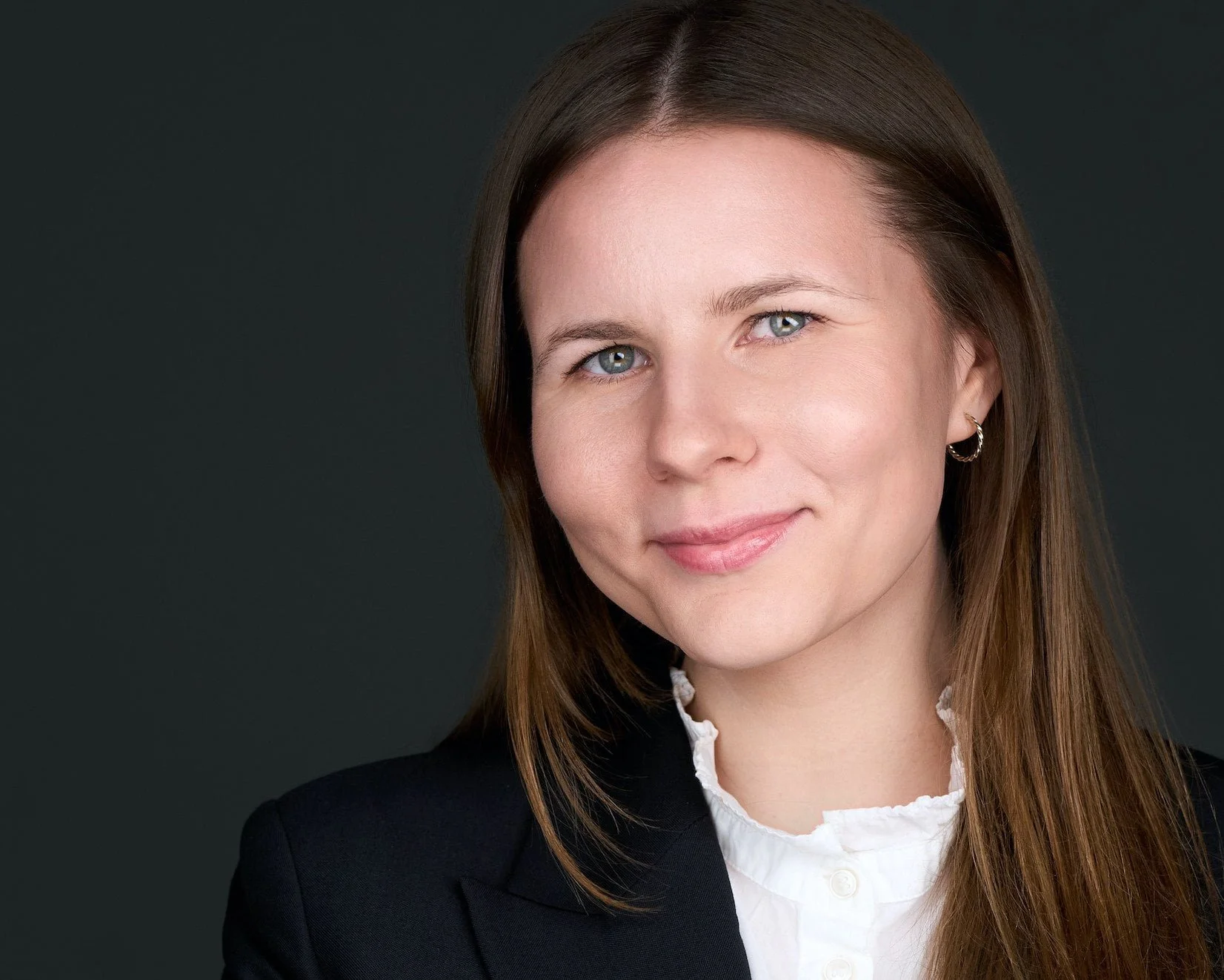 Close-up portrait of a woman with light skin, straight brown hair, blue eyes, wearing a black blazer and a white blouse, smiling softly against a dark background.