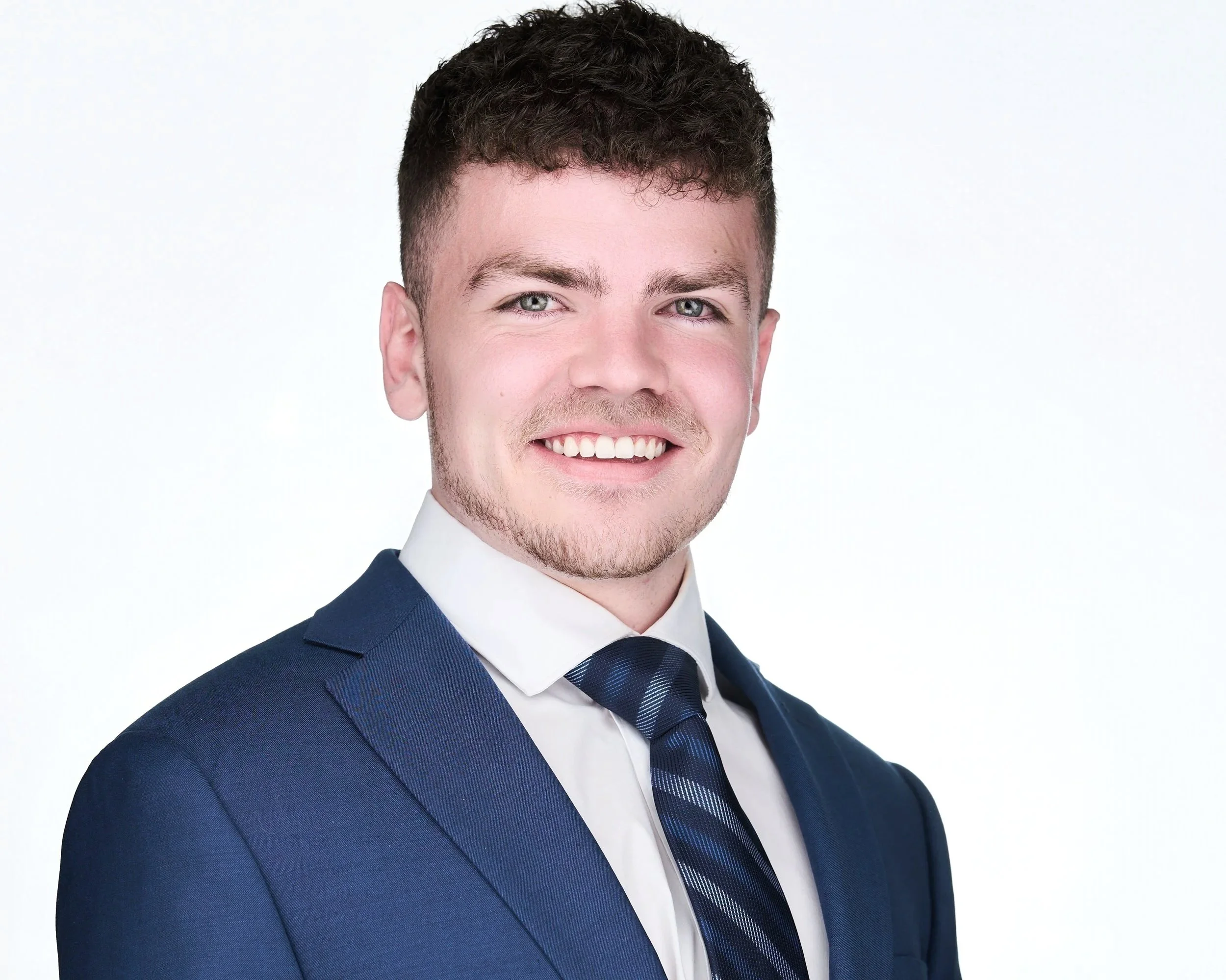 A young man in a dark blue suit and tie, smiling, against a plain white background.