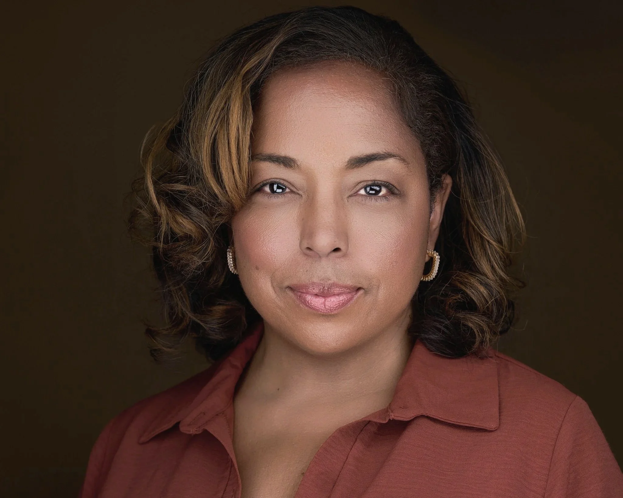 A woman with shoulder-length wavy hair, wearing gold earrings and a rust-colored shirt, looking directly at the camera with a neutral expression.