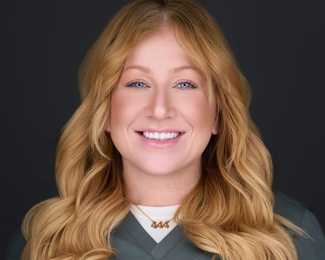 Close-up portrait of a smiling woman with long wavy red hair, blue eyes, and wearing a necklace with the number 444.