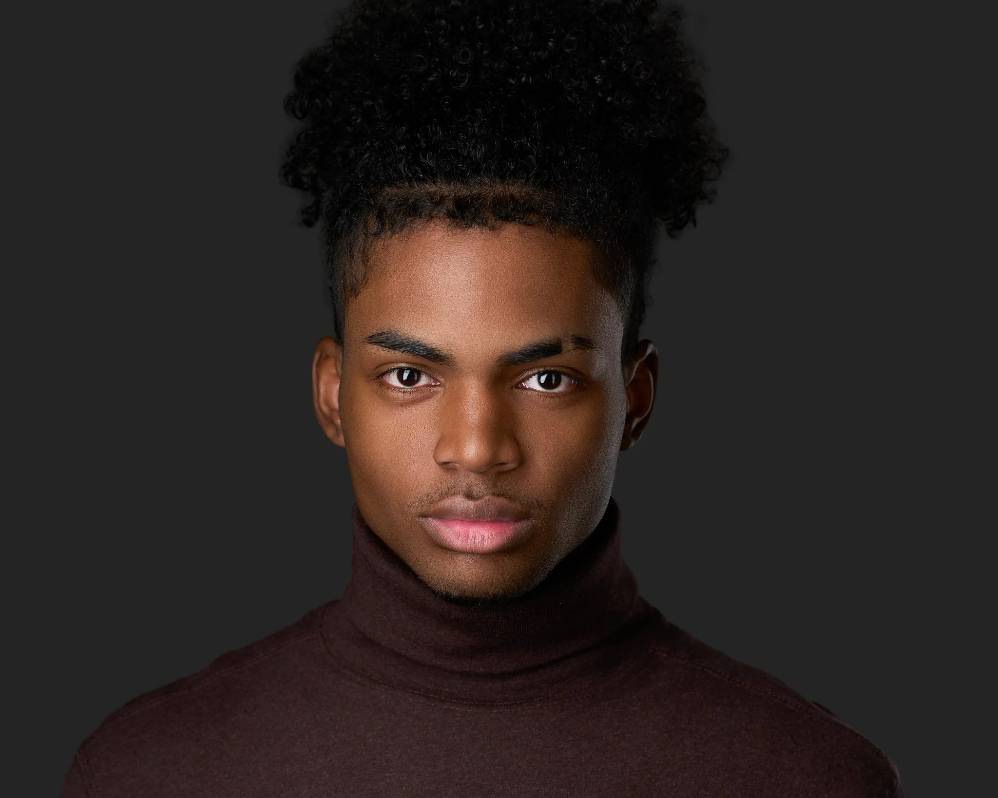 Professional studio headshot of a young man with with black hair pulled up, wearing a dark turtleneck, photographed against a clean dark gray background with soft, flattering lighting. Taken in Commerce City CO.