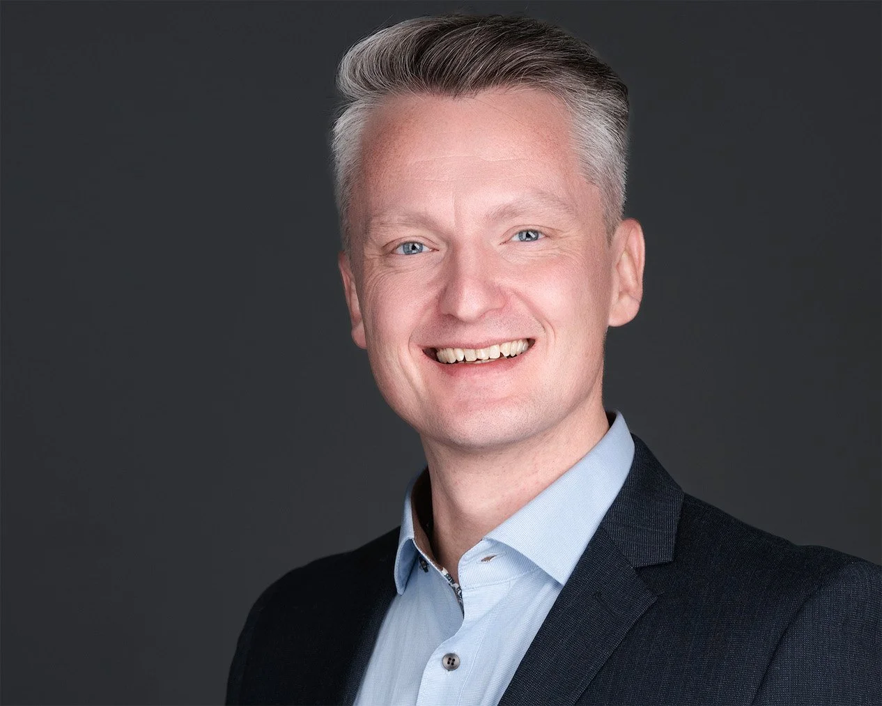Portrait of a man with short gray hair, blue eyes, smiling, wearing a dark suit jacket and light blue dress shirt, against a dark gray background.