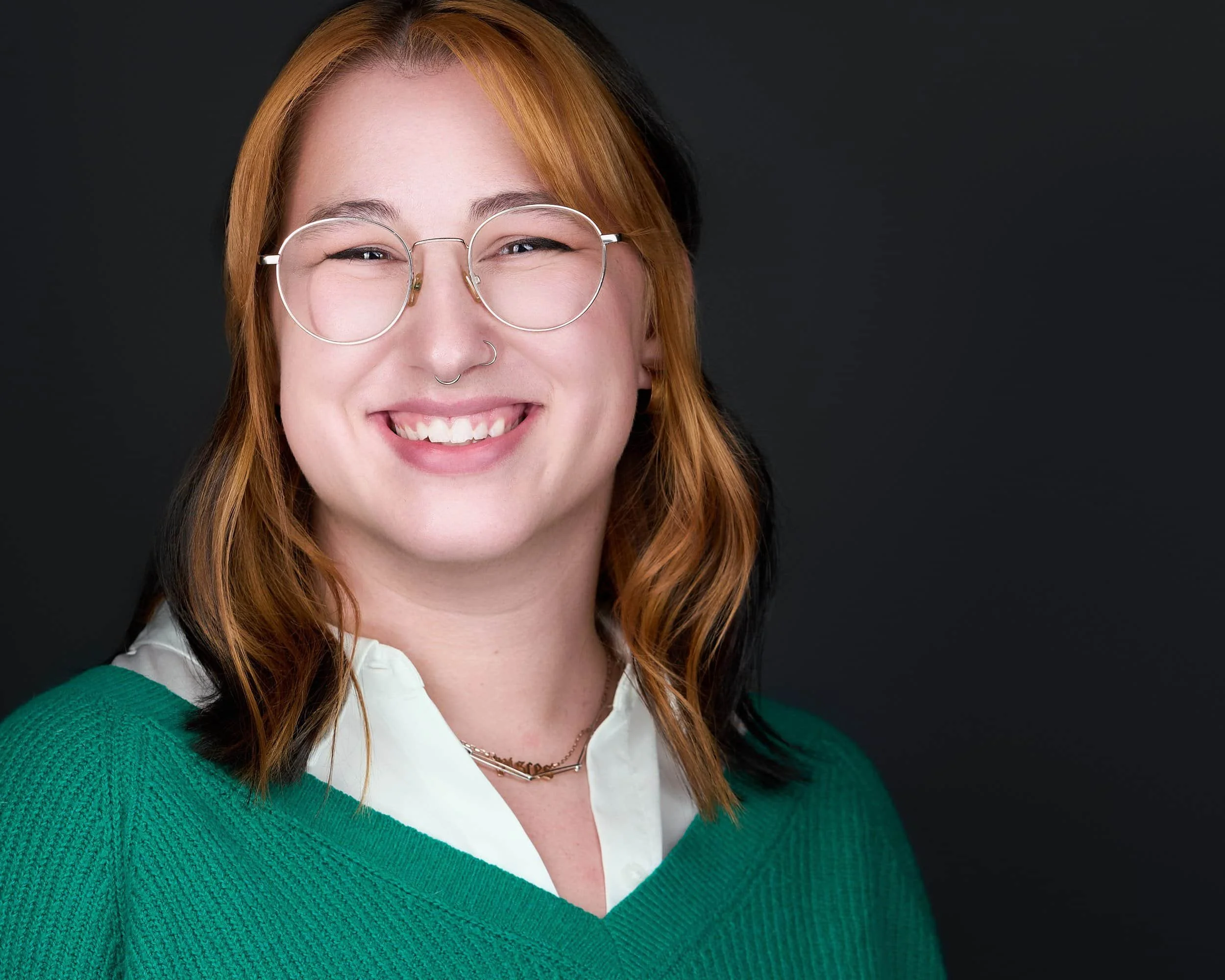 Portrait of smiling woman with glasses, septum piercing, white blouse, and green sweater against a dark background.