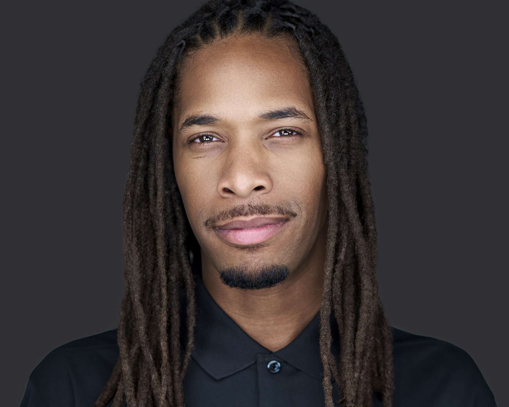 Close-up of a man with dreadlocks, wearing a black shirt, against a dark background.