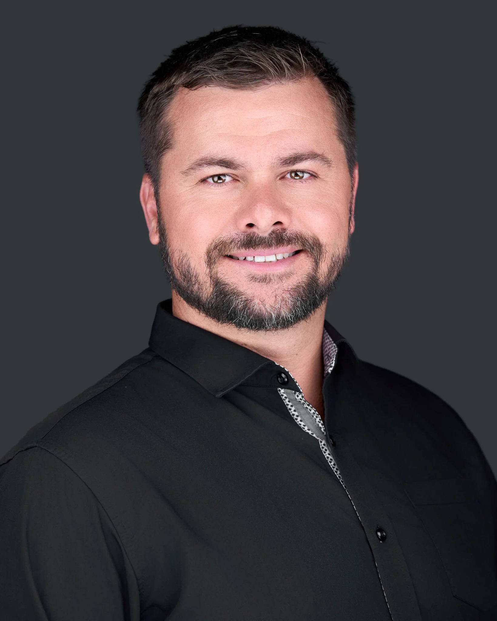 Portrait of a smiling man with short dark hair, beard, and mustache, wearing a black shirt with a patterned collar, against a dark gray background.